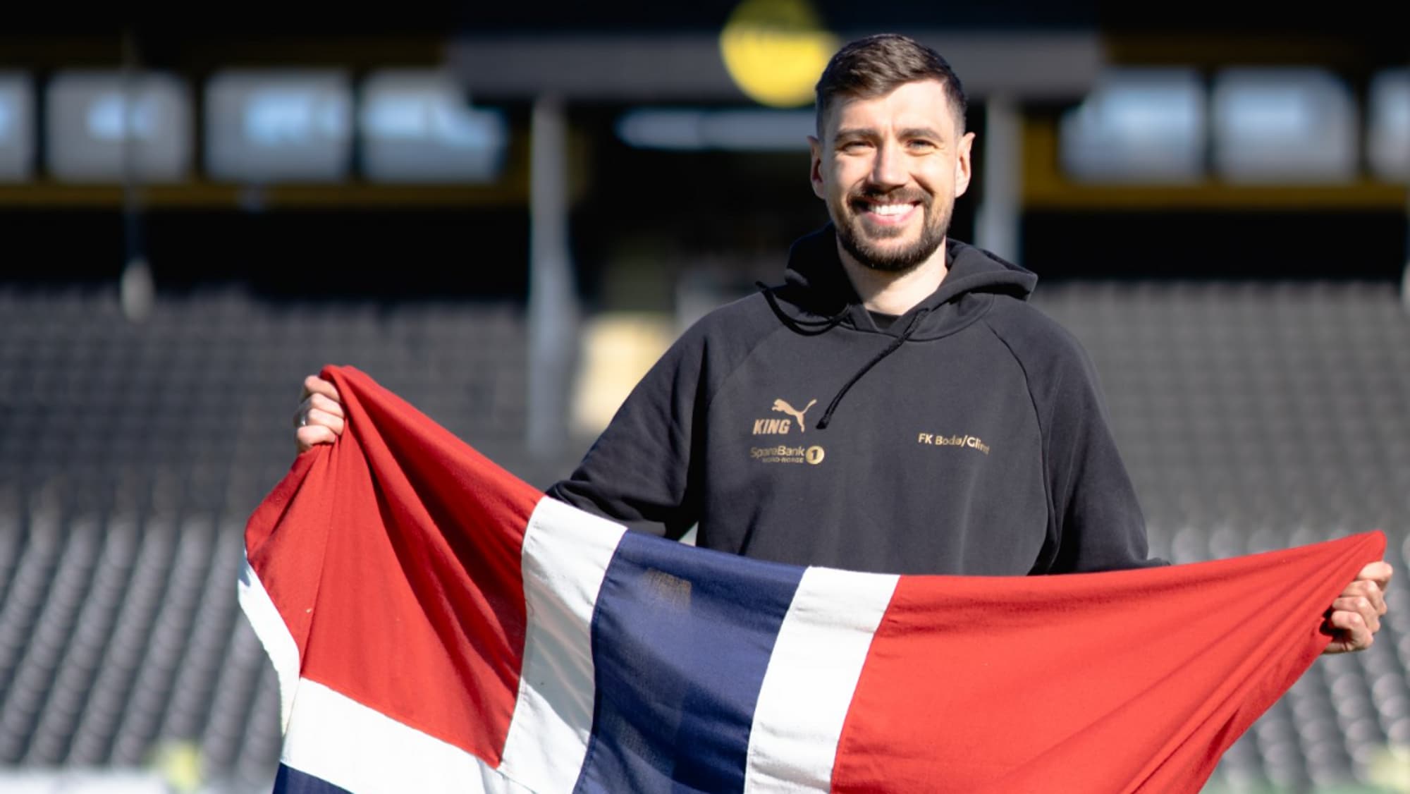 En mann med mørkt hår og skjegg som holder et norgeflagg på en fotballbane 