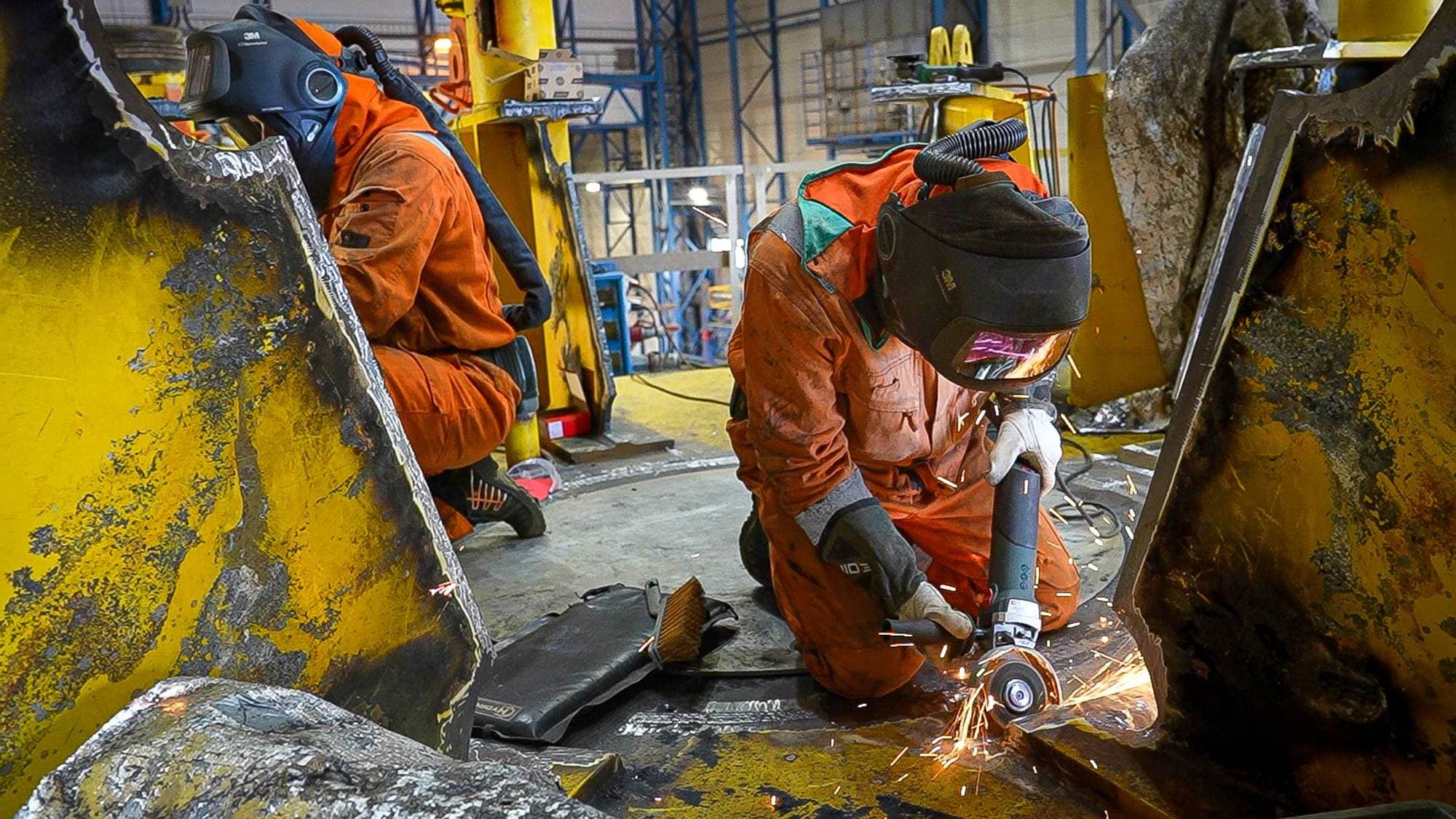 To industriarbeidere i oransje vernedresser og sveisehjelmer jobber med metall i et verksted. (Bildebeskrivelsen er laget av en KI-tjeneste)