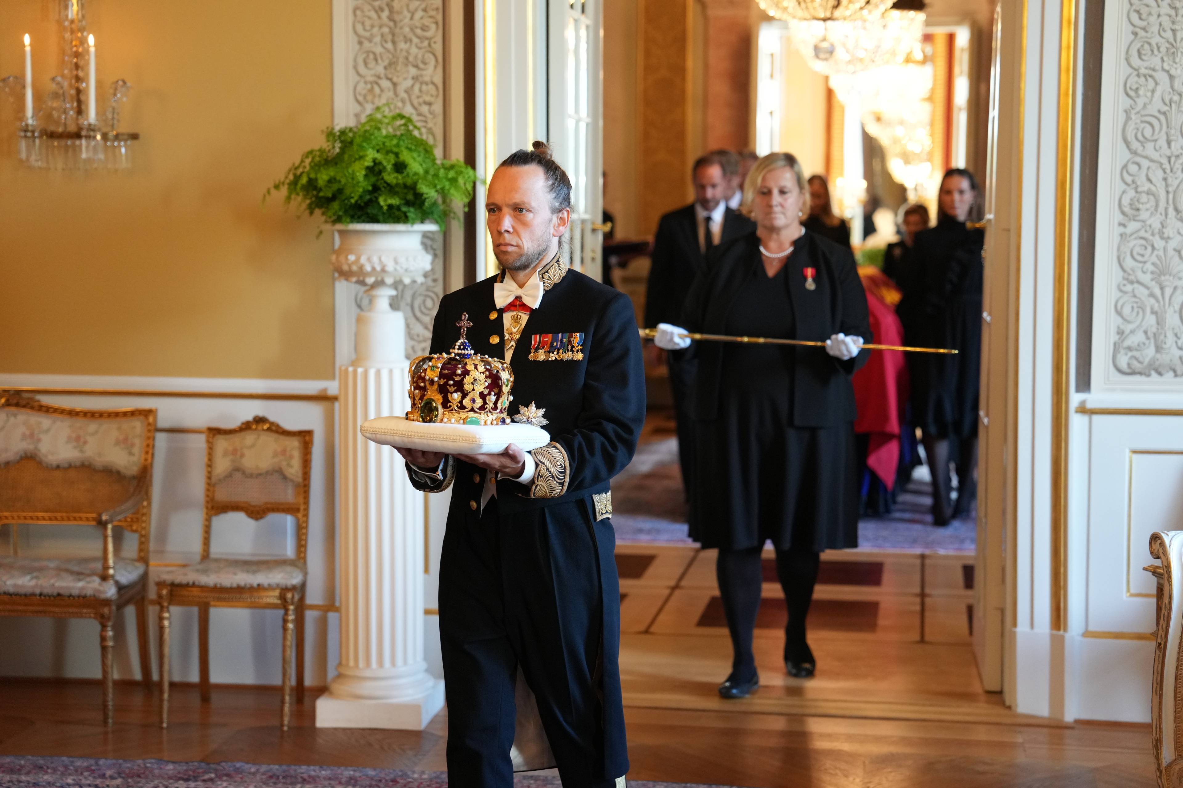 En mann i uniform bærer en krone på en pute i et ærverdig rom, fulgt av personer i svarte klær. (Bildebeskrivelsen er laget av en KI-tjeneste)