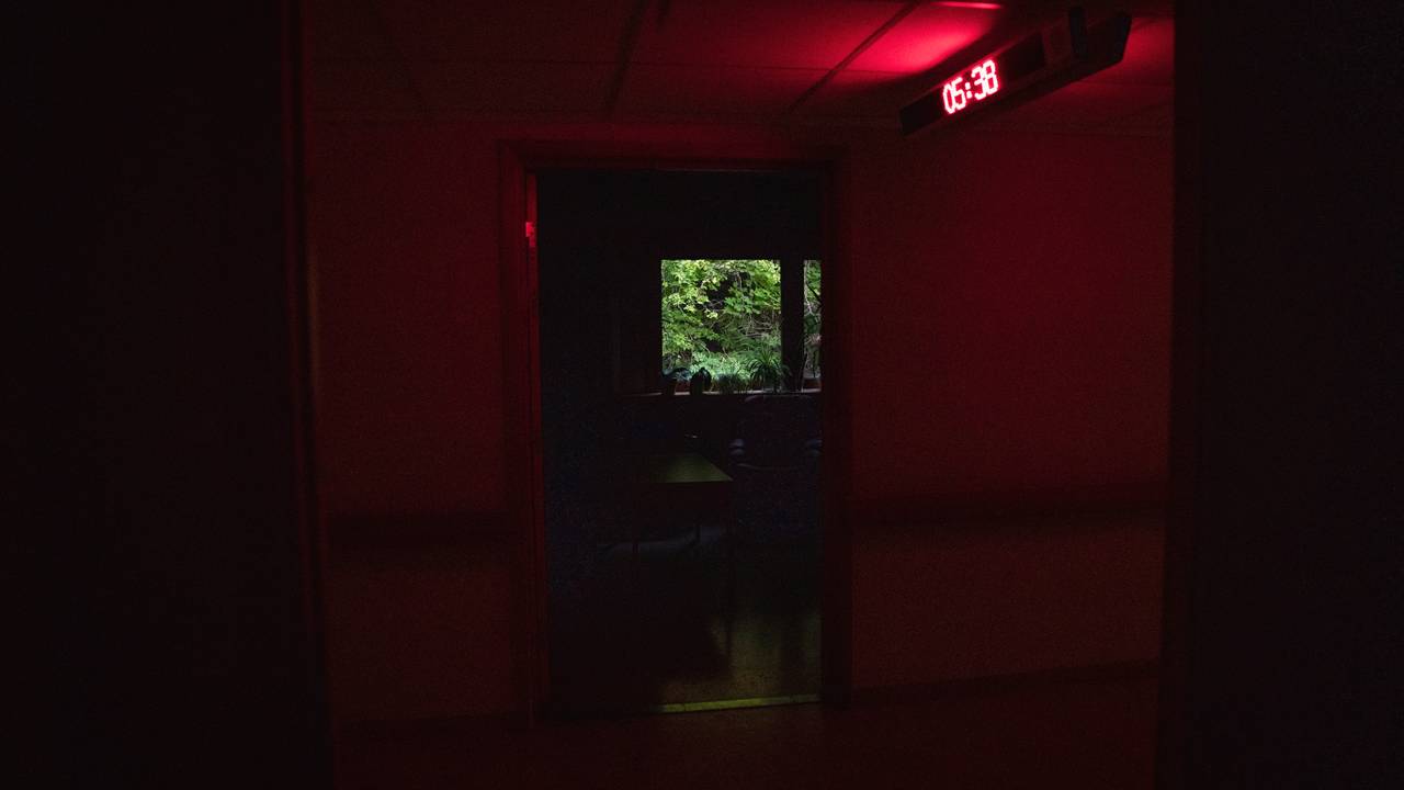 En mørk gang, kun opplyst av et rødt digitalt klokkeskilt i taket. I enden av et rom skimter man dagslyset som gryr. Den digitale klokken viser "05:38".