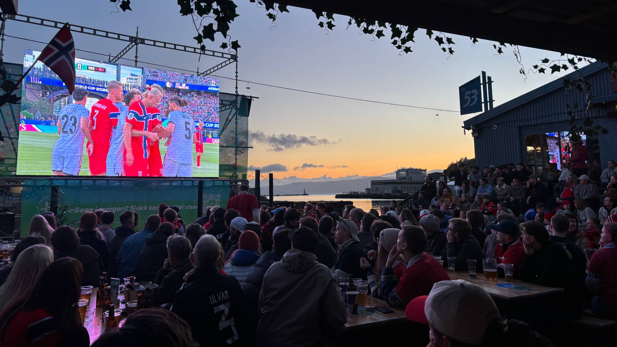 Folk ser fotballkamp på storskjerm utendørs ved sjøen i solnedgang. 