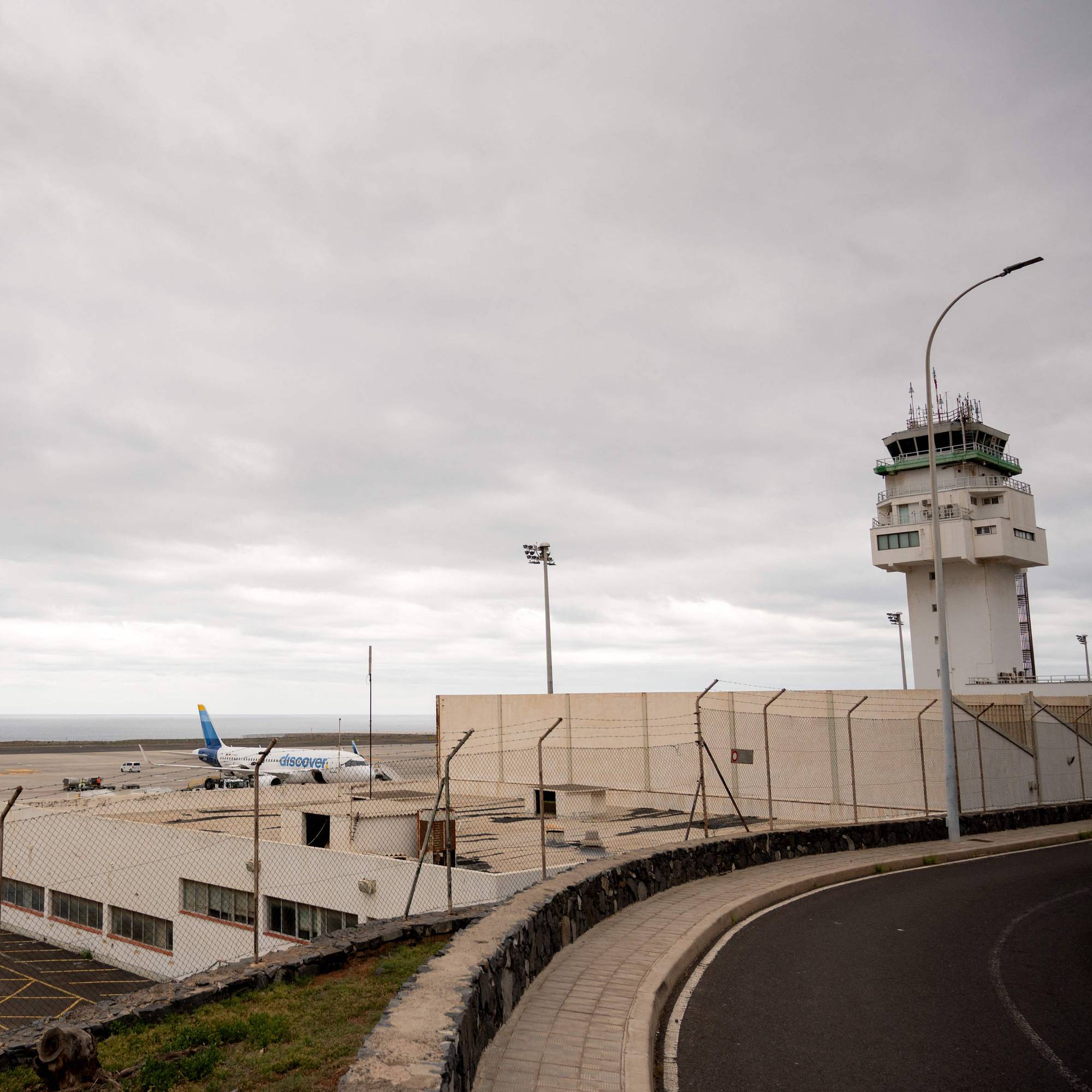 Et flyplasstårn står til høyre for bildet. Flere fly er parkert på rullebanen foran et hav. Veien til høyre er asfaltert. En klar himmel dekker bildet. (Bildebeskrivelsen er laget av en KI-tjeneste)