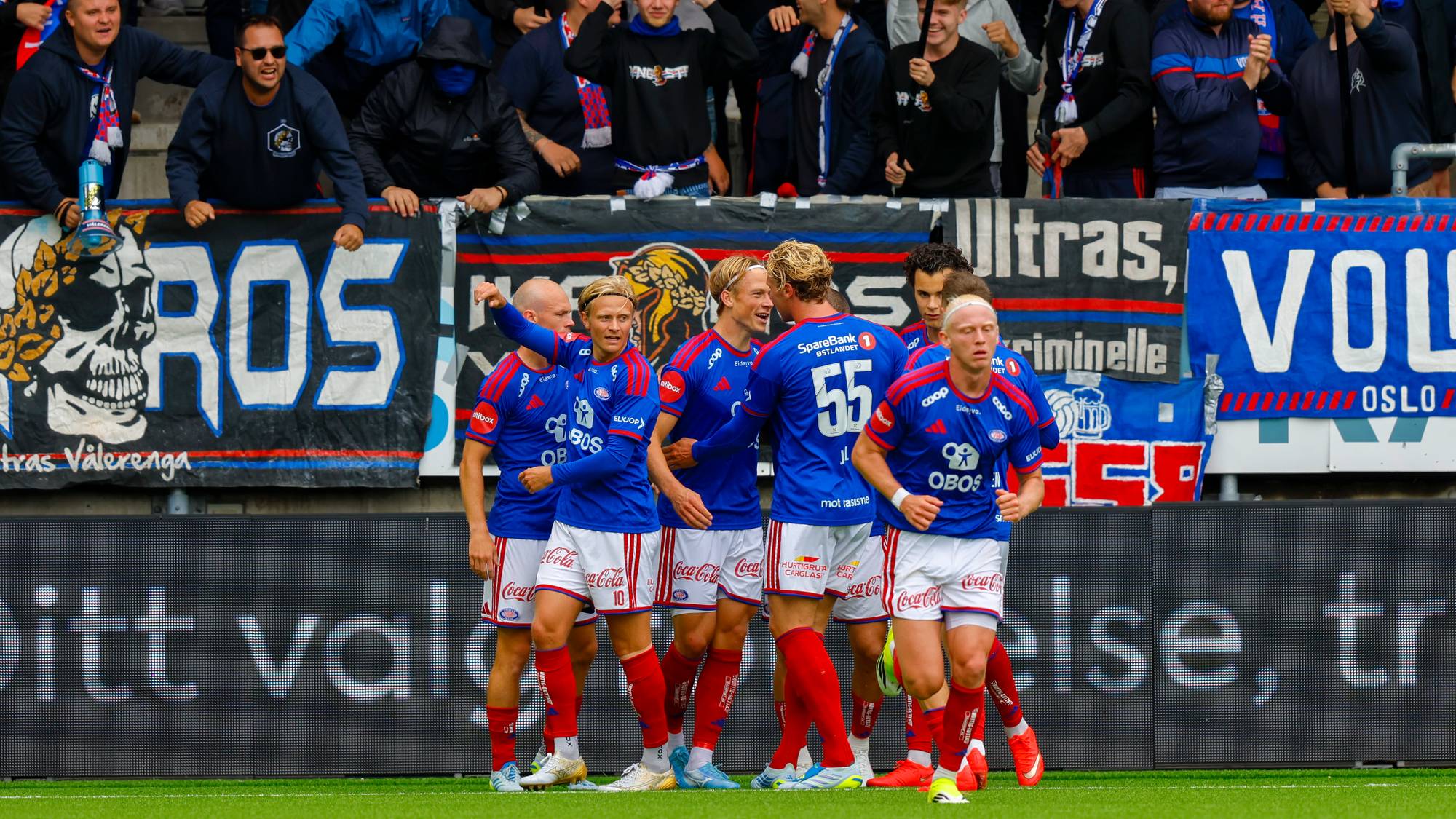 Fotballspillere i blå drakter jubler foran supportere på tribunen under en kamp. (Bildebeskrivelsen er laget av en KI-tjeneste)