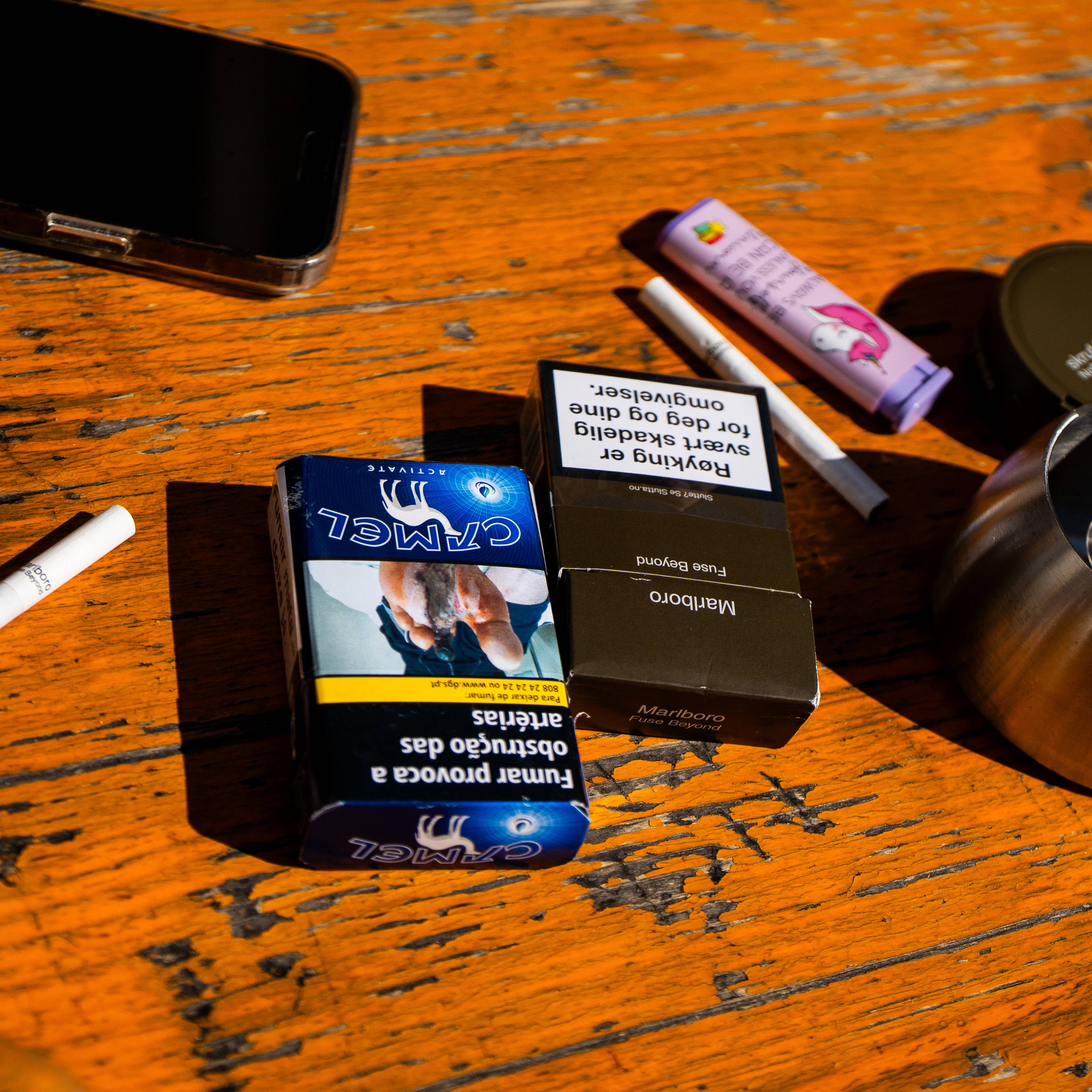 Bilde viser ein smarttelefon, ein pakke Camel-sigarettar og ein pakke Marlboro. Ved sidan av ligg det ein sigarett, ein leppepomade og et lite oskebeger. Alt ligg på et oransje trebord. (Bildebeskrivelsen er laget av ein KI-teneste)