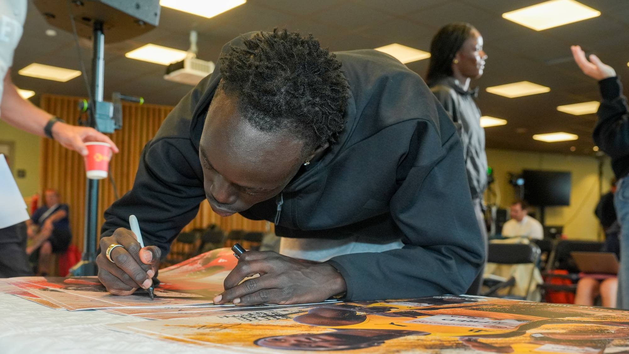 En mann med dreadlocks signerer et bilde. Han er ikledd en svart hettegenser og har en ring på fingeren. En person gir ham en kopp. I bakgrunnen er andre personer og en skjerm. (Bildebeskrivelsen er laget av en KI-tjeneste)