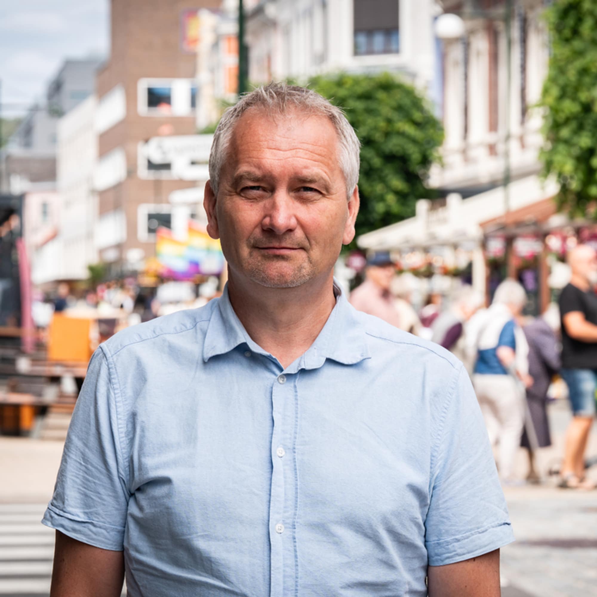 En mann med kort, grått hår og blå skjorte står i en gate. I bakgrunnen ser man mange folk.