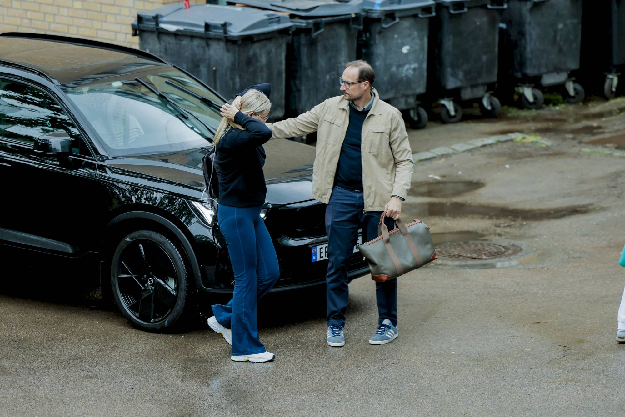 Kronprinsparet står ved en bil. De er i ferd med å sette seg inn i bilen.Bak dem står det flere søppelcontainere