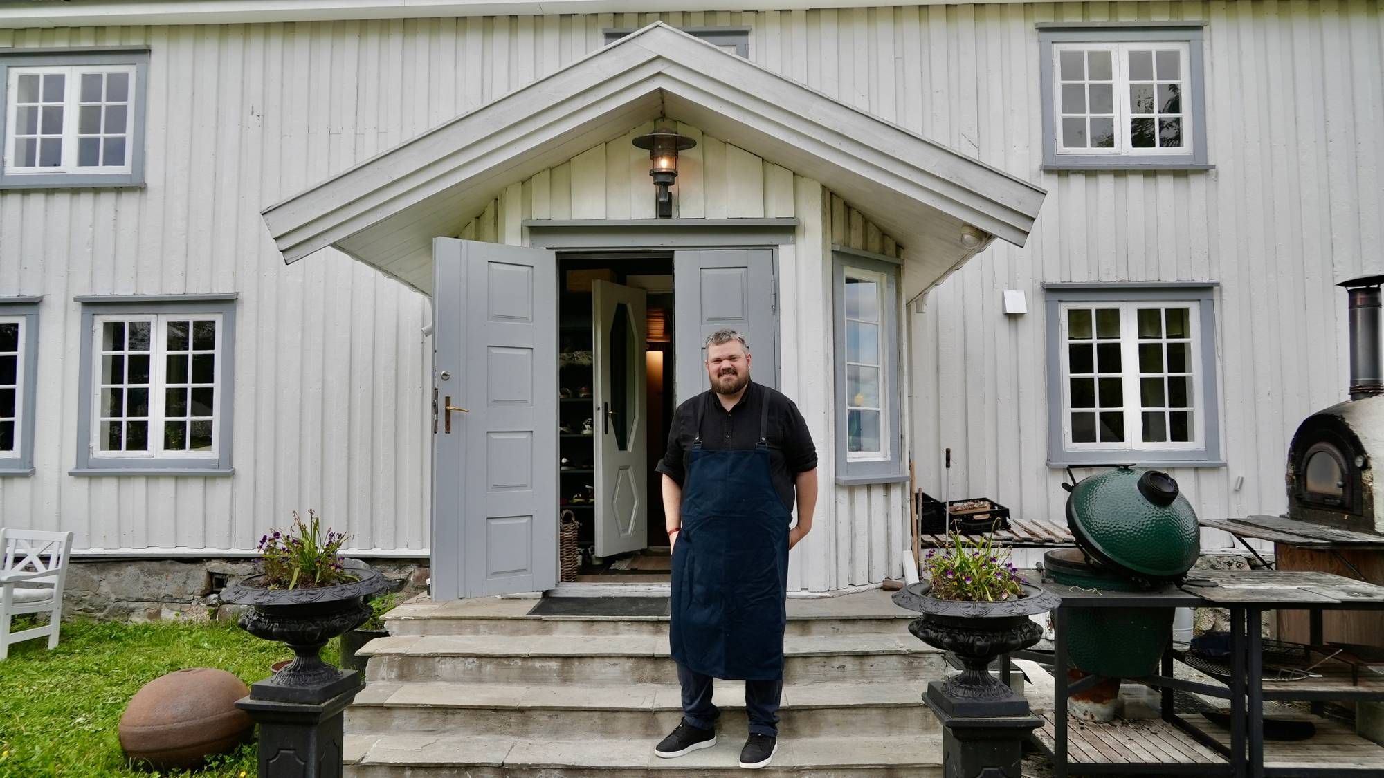En mann i kokkeklær står på trappen foran en hvit bygning. Mannen smiler mot kameraet. Bygningen har grå dører og vinduer. Utenfor står det en ovn og en kullgrill. (Bildebeskrivelsen er laget av en KI-tjeneste)
