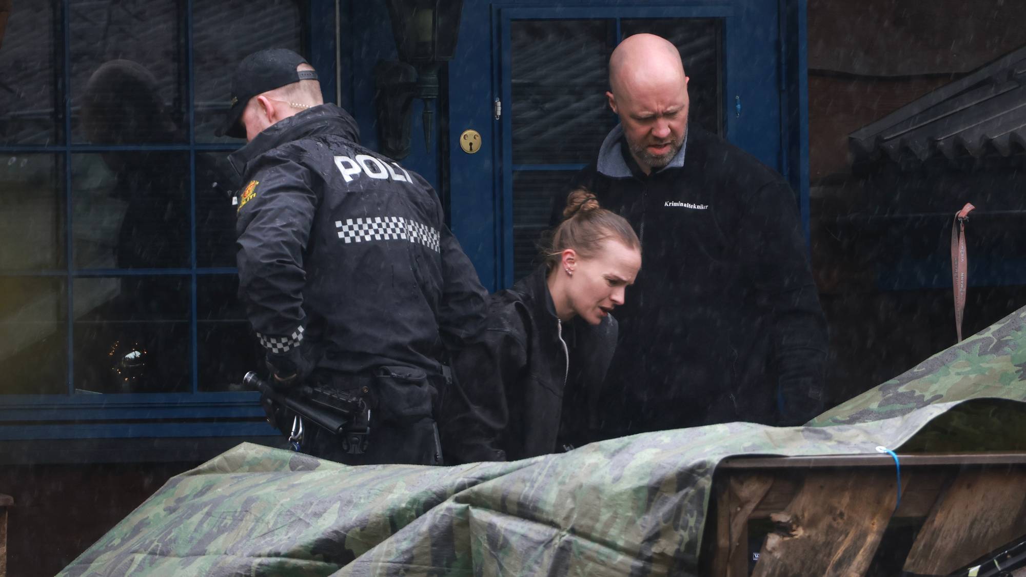 Politifolk er på stedet. En kvinne er på vei ut av en blå dør. Det faller regn. En presenning dekker noe foran. (Bildebeskrivelsen er laget av en KI-tjeneste)