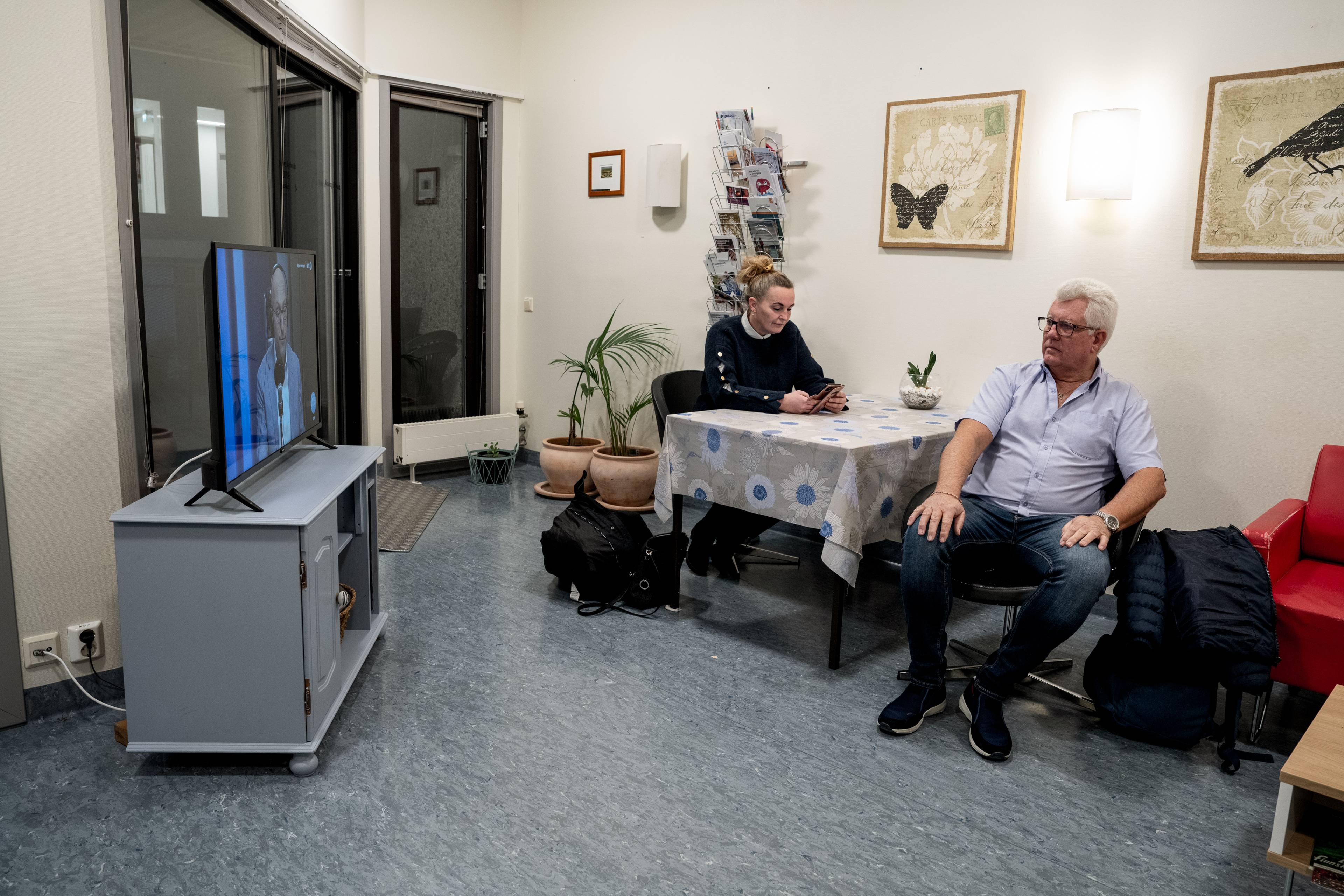 Erik på venterommet på sykehuset med datteren Camilla.  De ser på tv og sitter ved et bord med plastduk. Bak dem henger to bilder.