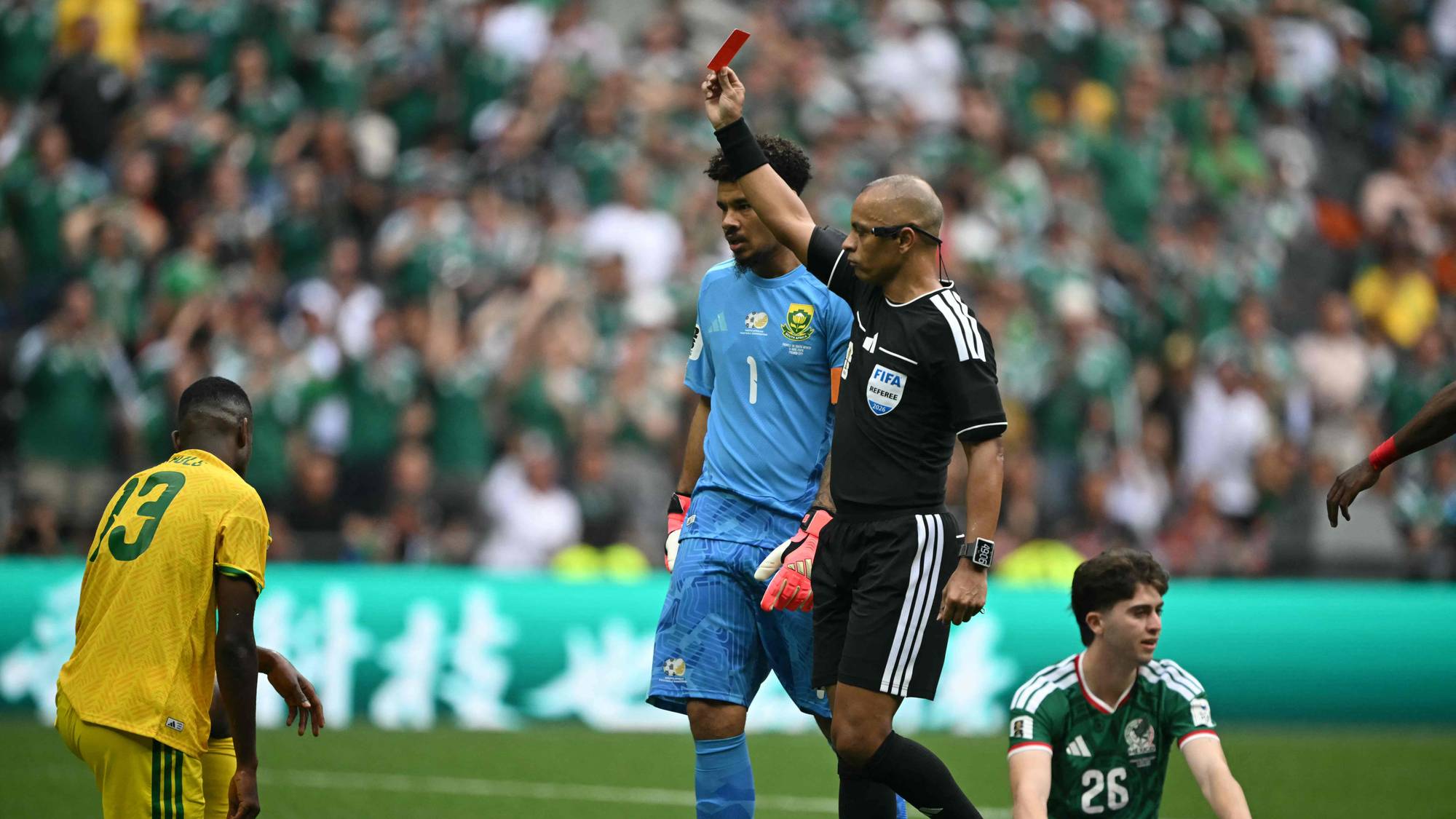 Dommer viser rødt kort til keeper. En annen spiller med nummer 13 sitter på kne på gressmatten. En spiller med nummer 26 sitter på bakken. Publikum ser på i bakgrunnen. (Bildebeskrivelsen er laget av en KI-tjeneste)