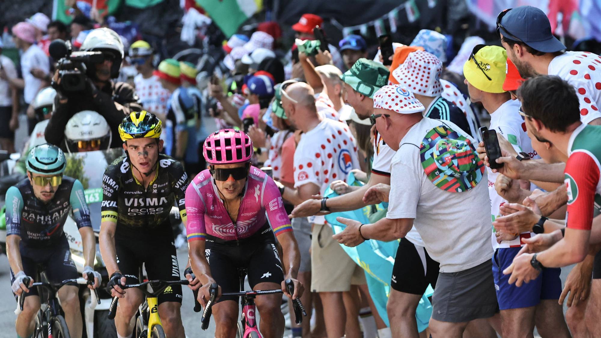 Syklister i Tour de France sykler forbi publikum langs veien. Flere flagg er synlige i bakgrunnen. (Bildebeskrivelsen er laget av en KI-tjeneste)