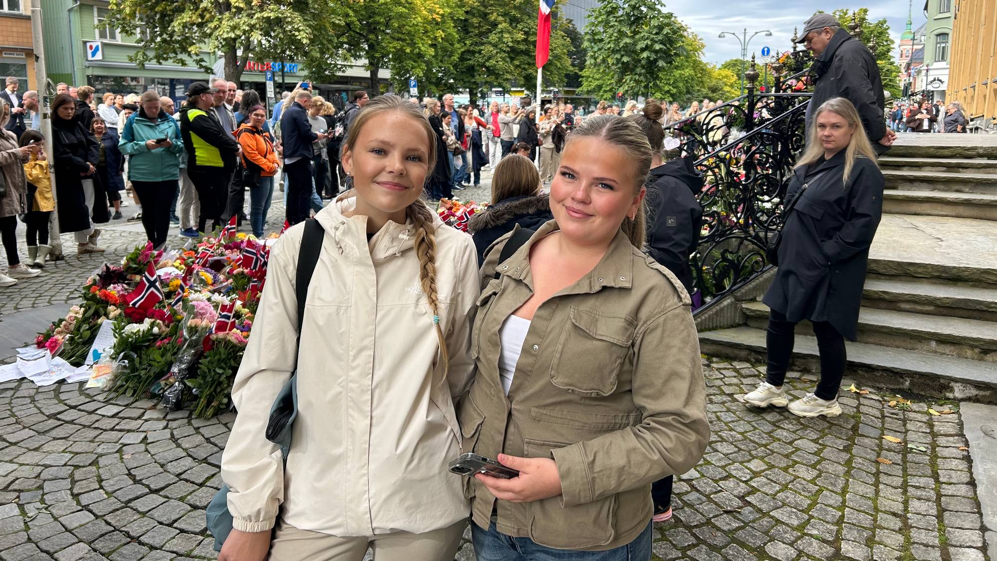 To unge kvinner står foran et blomsterhav ved en minnesmarkering i en bygate. 