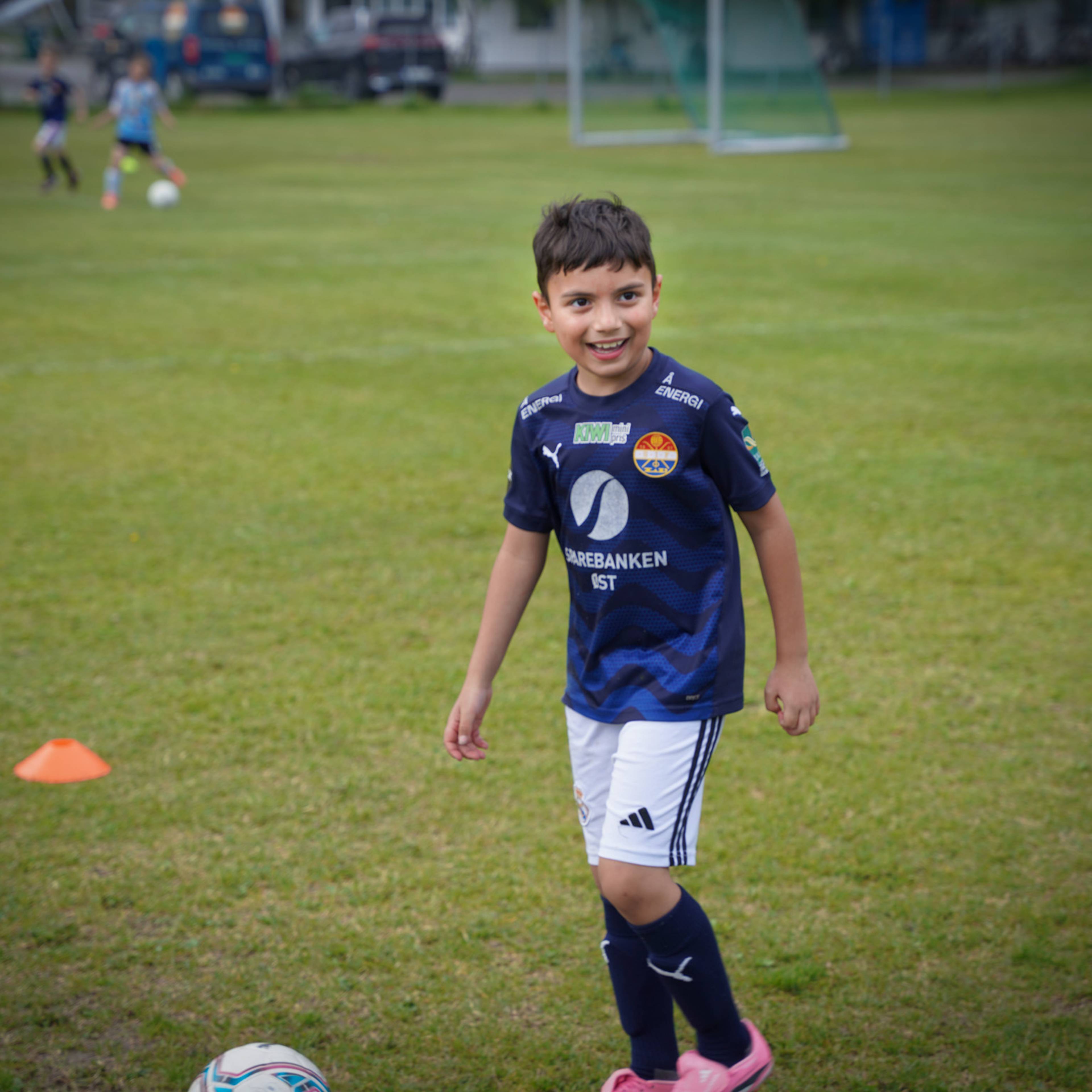 En gutt i fotballdrakt ser mot kameraet på en gressbane. Han smiler og står i en treningssituasjon. Bak ham er andre barn som spiller fotball. (Bildebeskrivelsen er laget av en KI-tjeneste)
