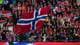 NTB En stor folkemengde er samlet på en idrettsarena og vifter med norske flagg og supporterutstyr. Mange av tilskuerne har på seg supporterutstyr med norske farger og symboler. Scenen er dekket av norske flagg, inkludert et stort flagg som dominerer forgrunnen. Stemningen er preget av patriotisme og støtte til det norske landslaget. (Bildebeskrivelsen er laget av en KI-tjeneste)