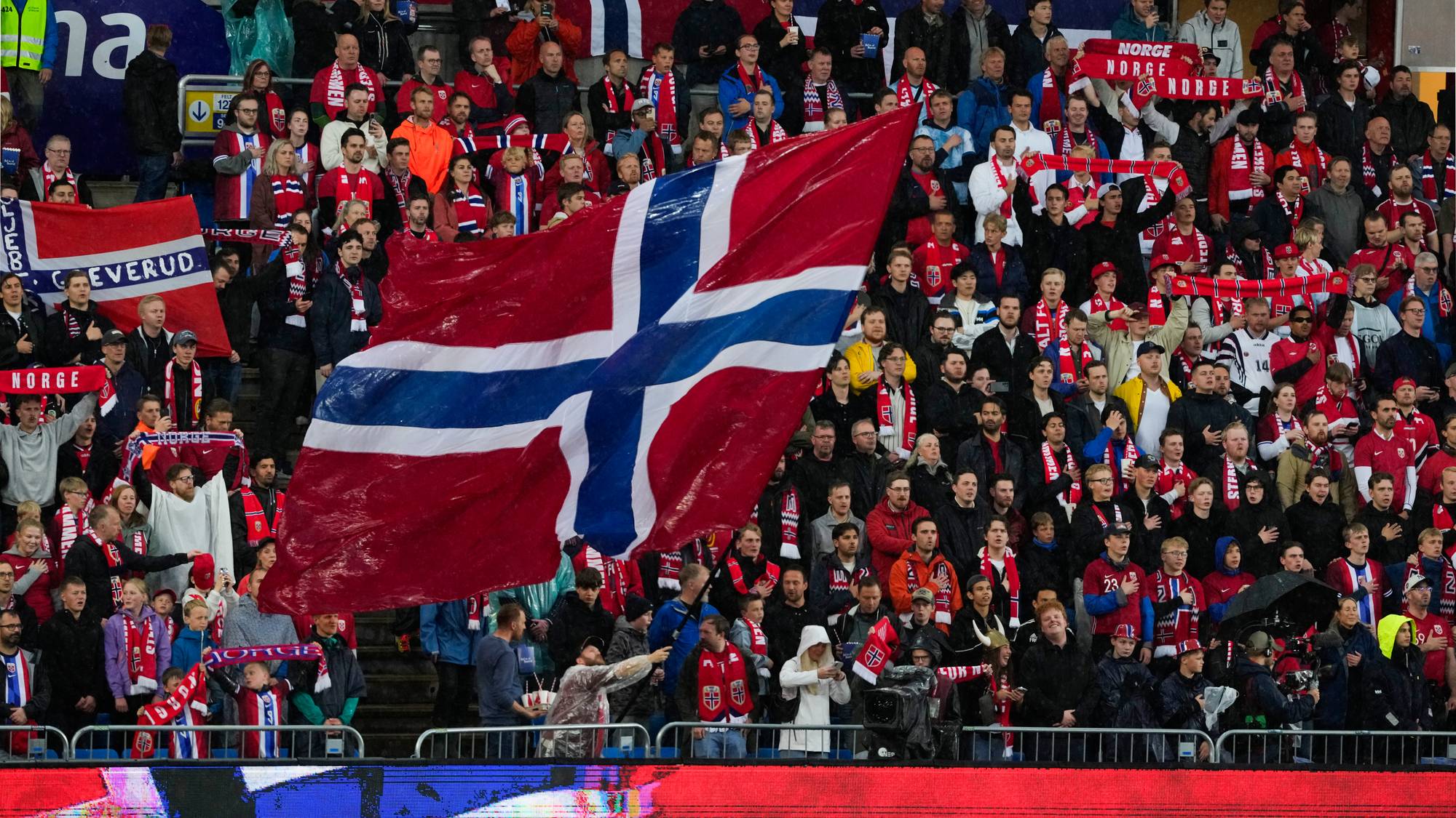 En stor folkemengde er samlet på en idrettsarena og vifter med norske flagg og supporterutstyr. Mange av tilskuerne har på seg supporterutstyr med norske farger og symboler. Scenen er dekket av norske flagg, inkludert et stort flagg som dominerer forgrunnen. Stemningen er preget av patriotisme og støtte til det norske landslaget. (Bildebeskrivelsen er laget av en KI-tjeneste)