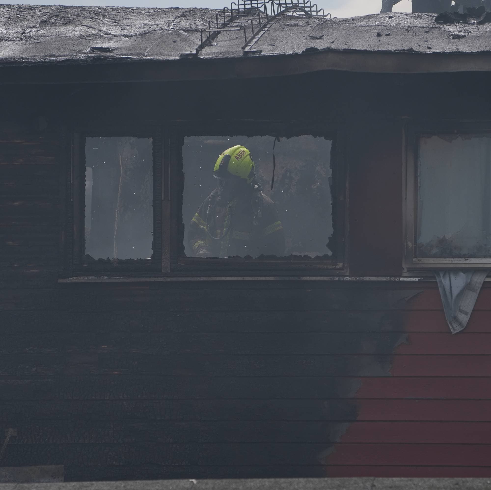 En brannmann med gul hjelm og oransje bekledning kikker ut av et vindu i et hus som har brent. Huset er røyk- og sot-skadet. Taket ser ut til å ha delvis kollapset. (Bildebeskrivelsen er laget av en KI-tjeneste)