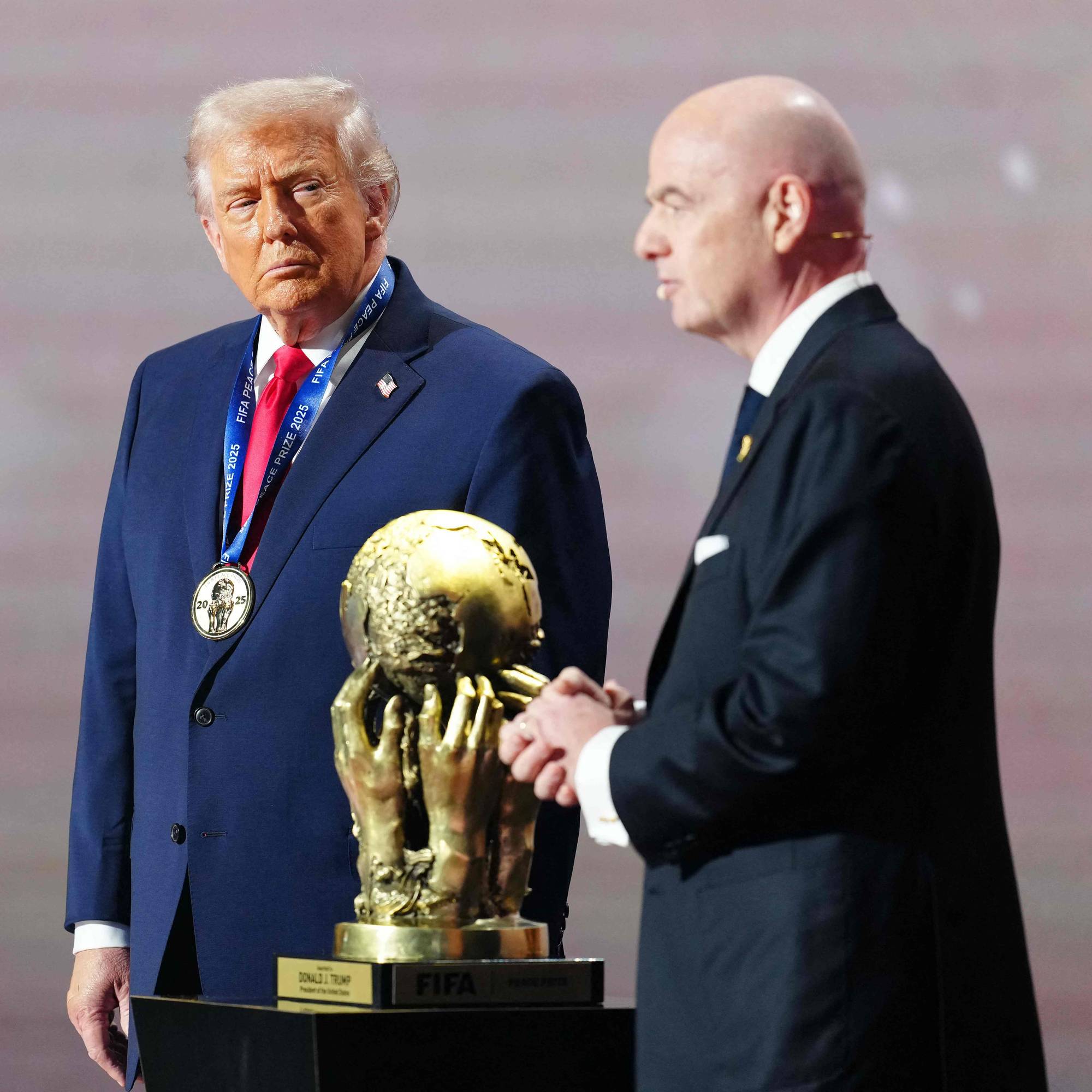 Donald Trump og Gianni Infantino står ved siden av en premie. Trump har en medalje rundt halsen. Prisen er en gullklode støttet av hender. (Bildebeskrivelsen er laget av en KI-tjeneste)