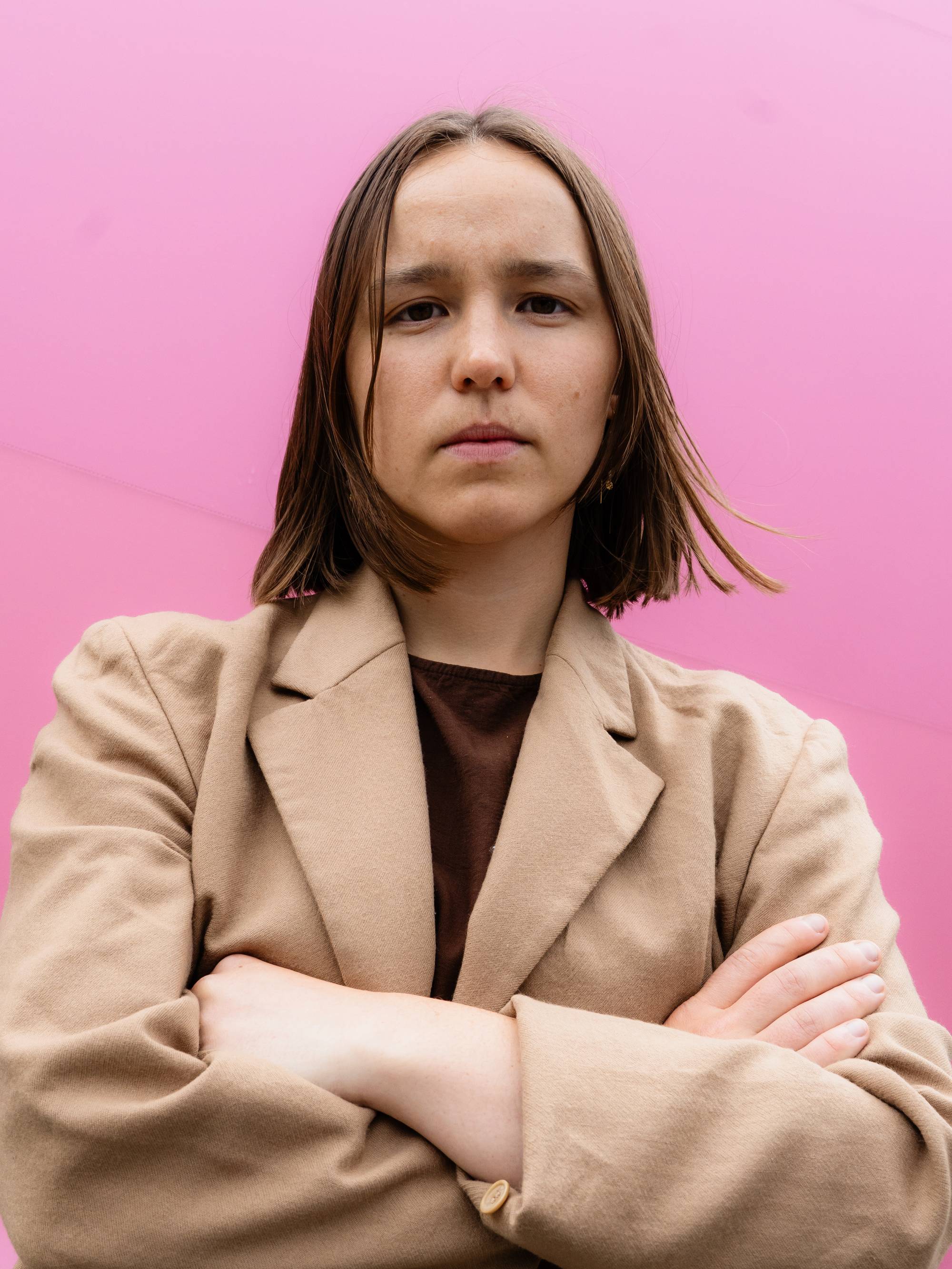 Portrett av ung kvinne med alvorlig uttrykkiansiktet og armene i kors. Rosa bakgrunn.