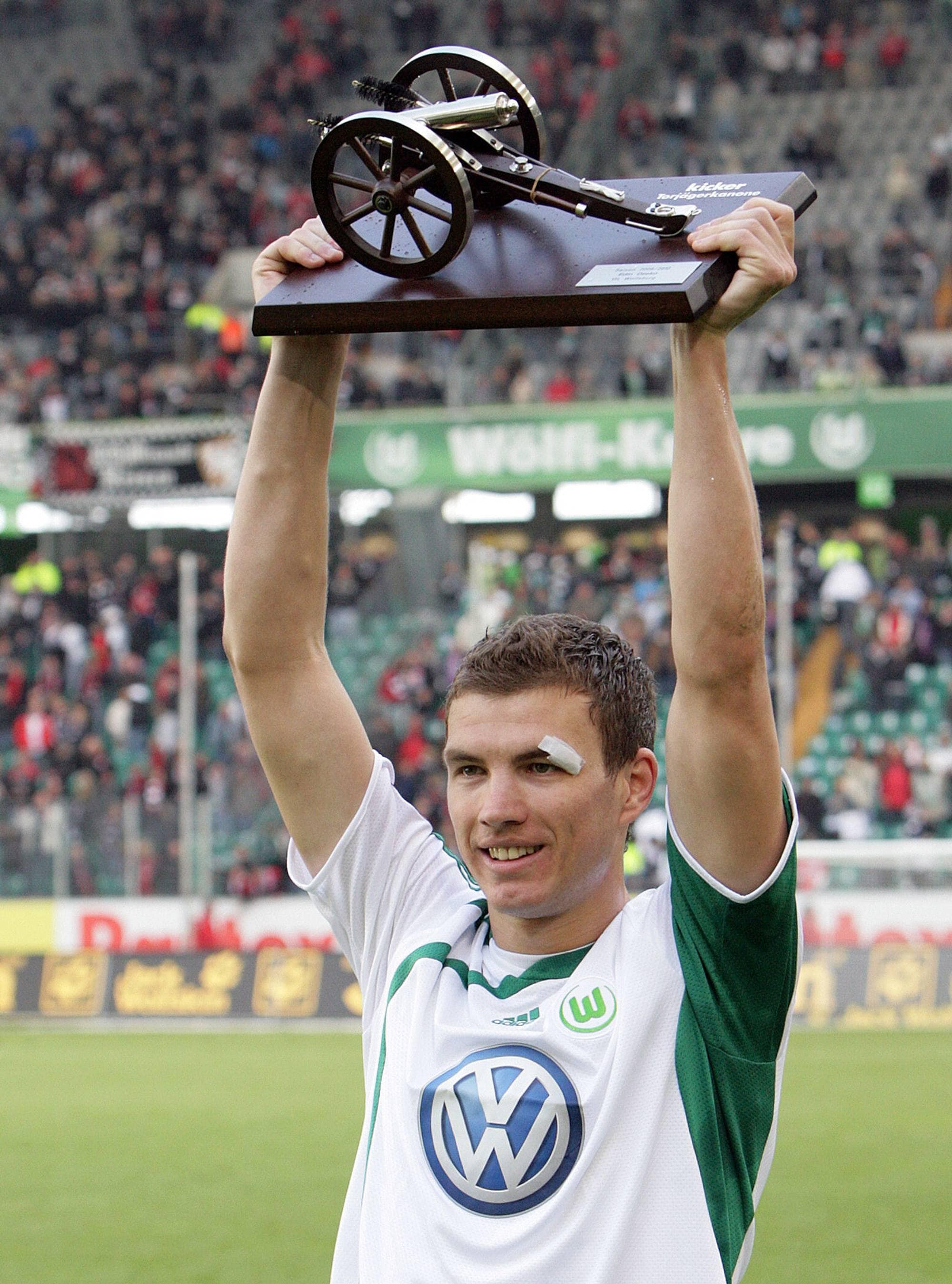 En fotballspiller holder en pokal med en kanon i tre. Han er iført en hvit trøye med grønn krage og V-formet logo. Bak ham er et publikum på en stadion. (Bildebeskrivelsen er laget av en KI-tjeneste)