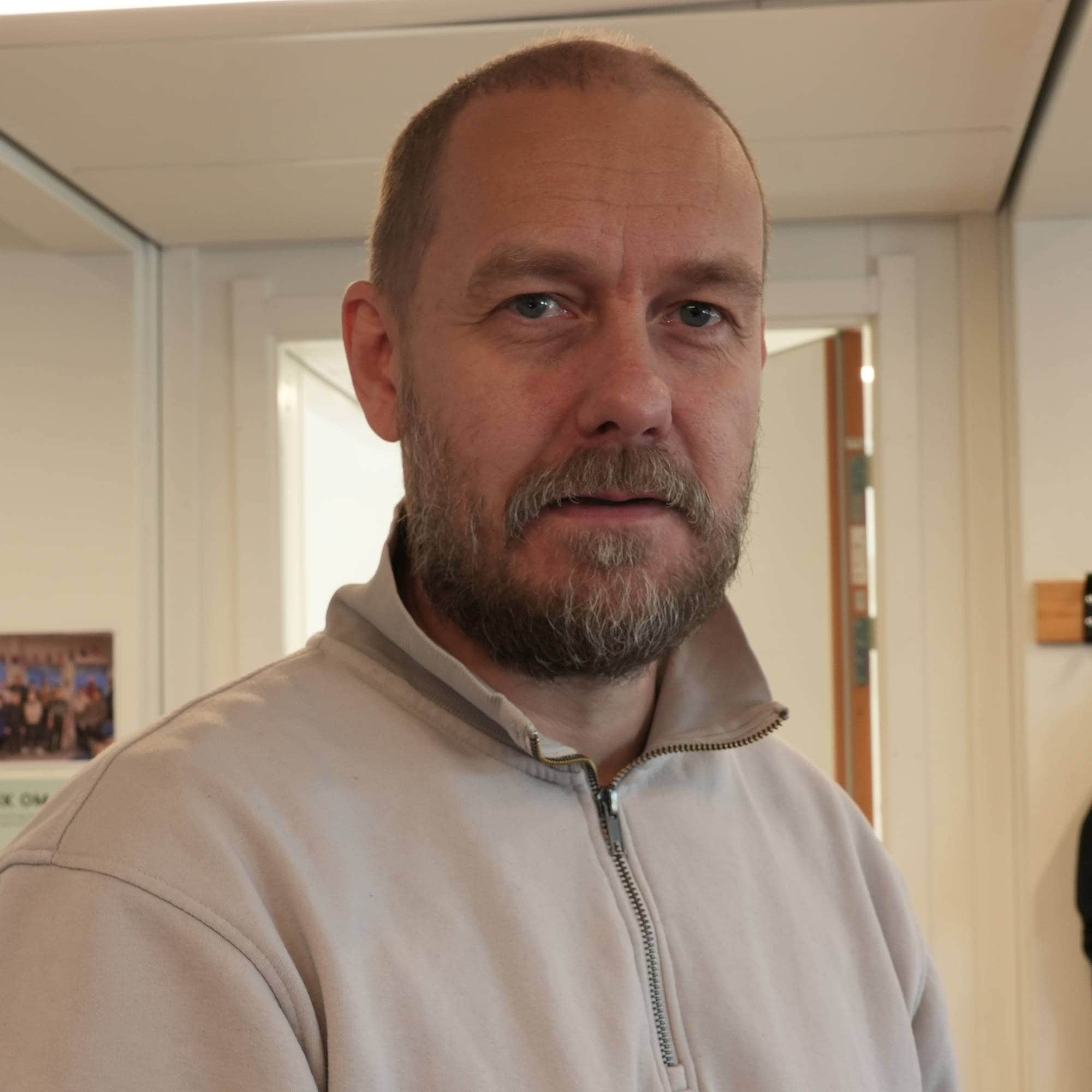Mann med skjegg i lys genser står i et klasserom med plakater på veggen. (Bildebeskrivelsen er laget av en KI-tjeneste)