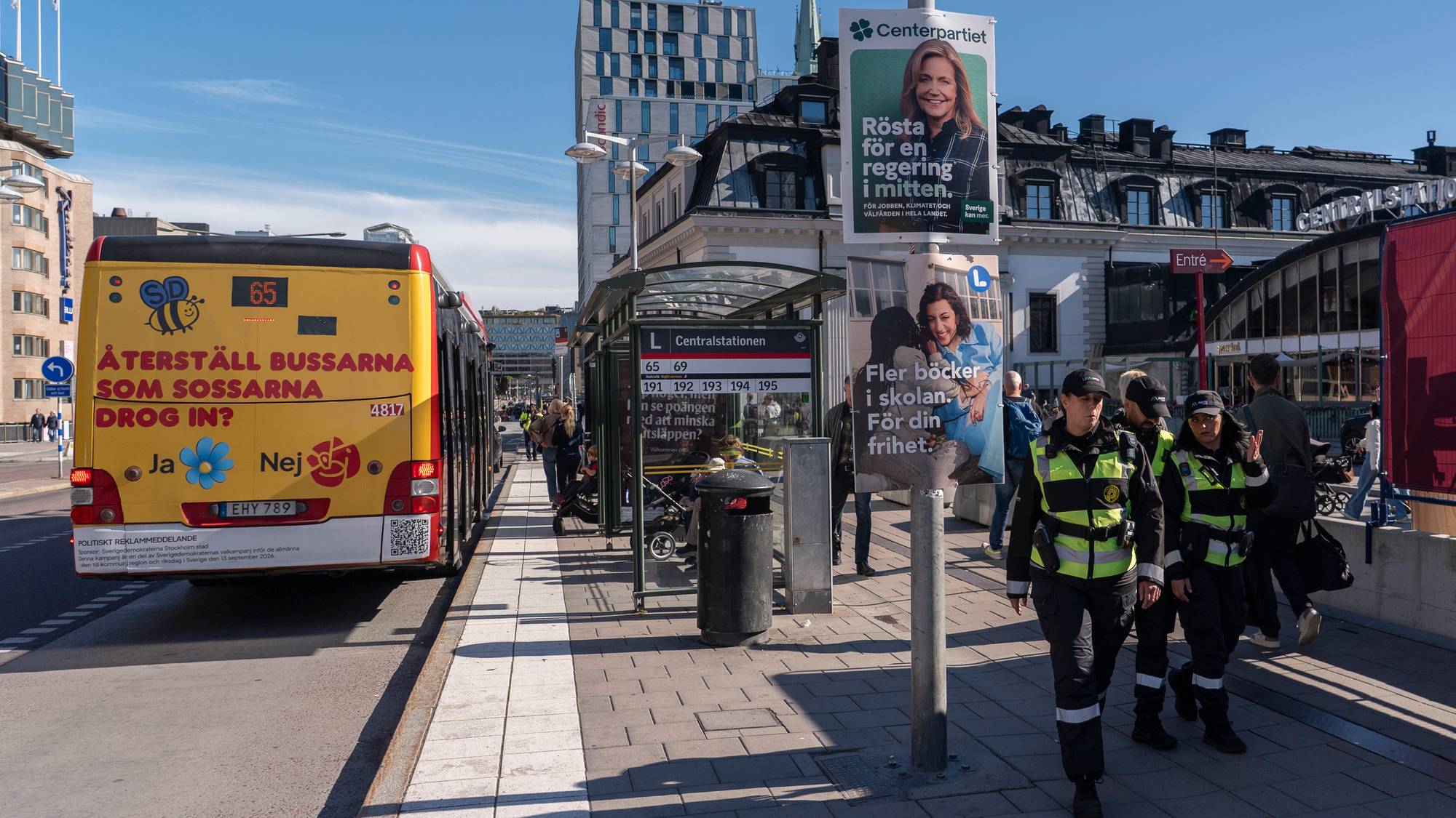 Vektere går forbi valgplakater ved en bussholdeplass i Stockholm, mens en buss med valgbudskap står. (Bildebeskrivelsen er laget av en KI-tjeneste)