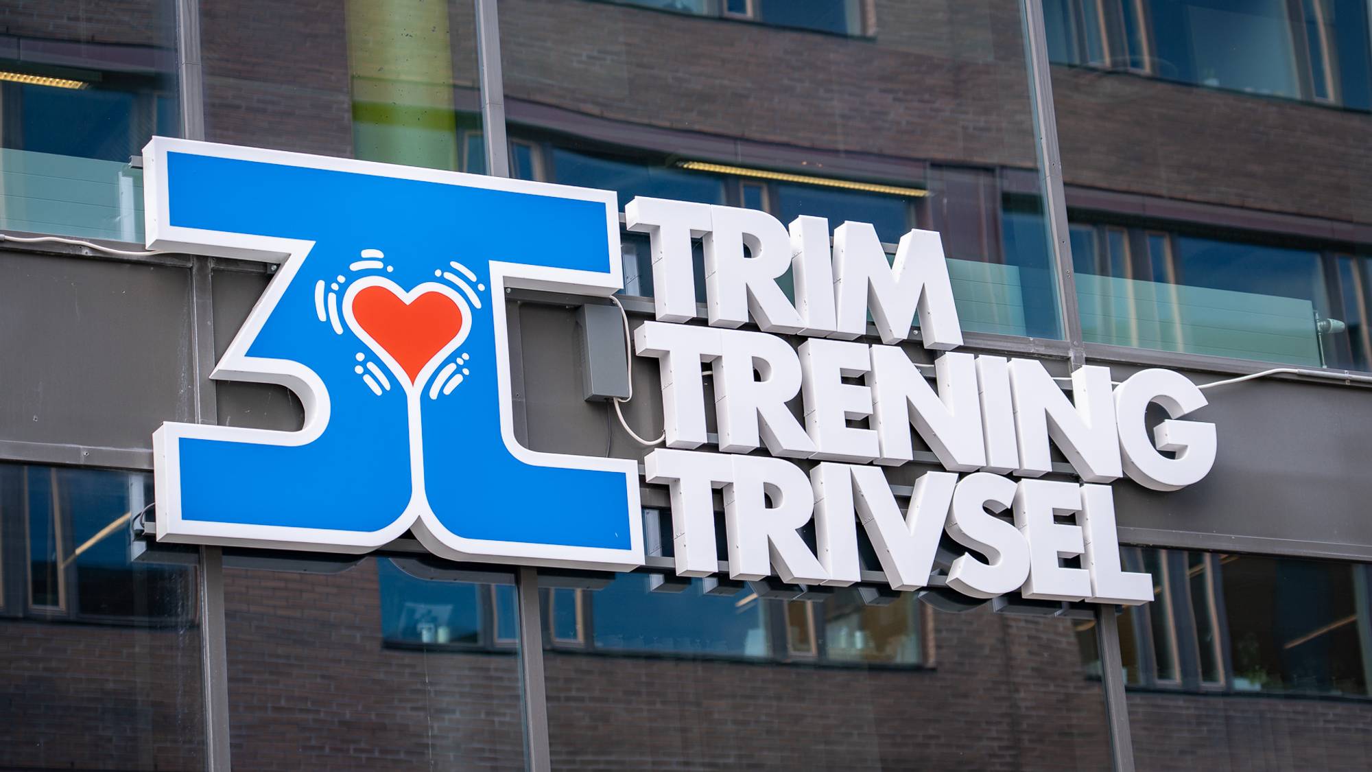 Logoen "3T Trim Trening Trivsel" henger på en glassvegg på utsiden av et bygg.