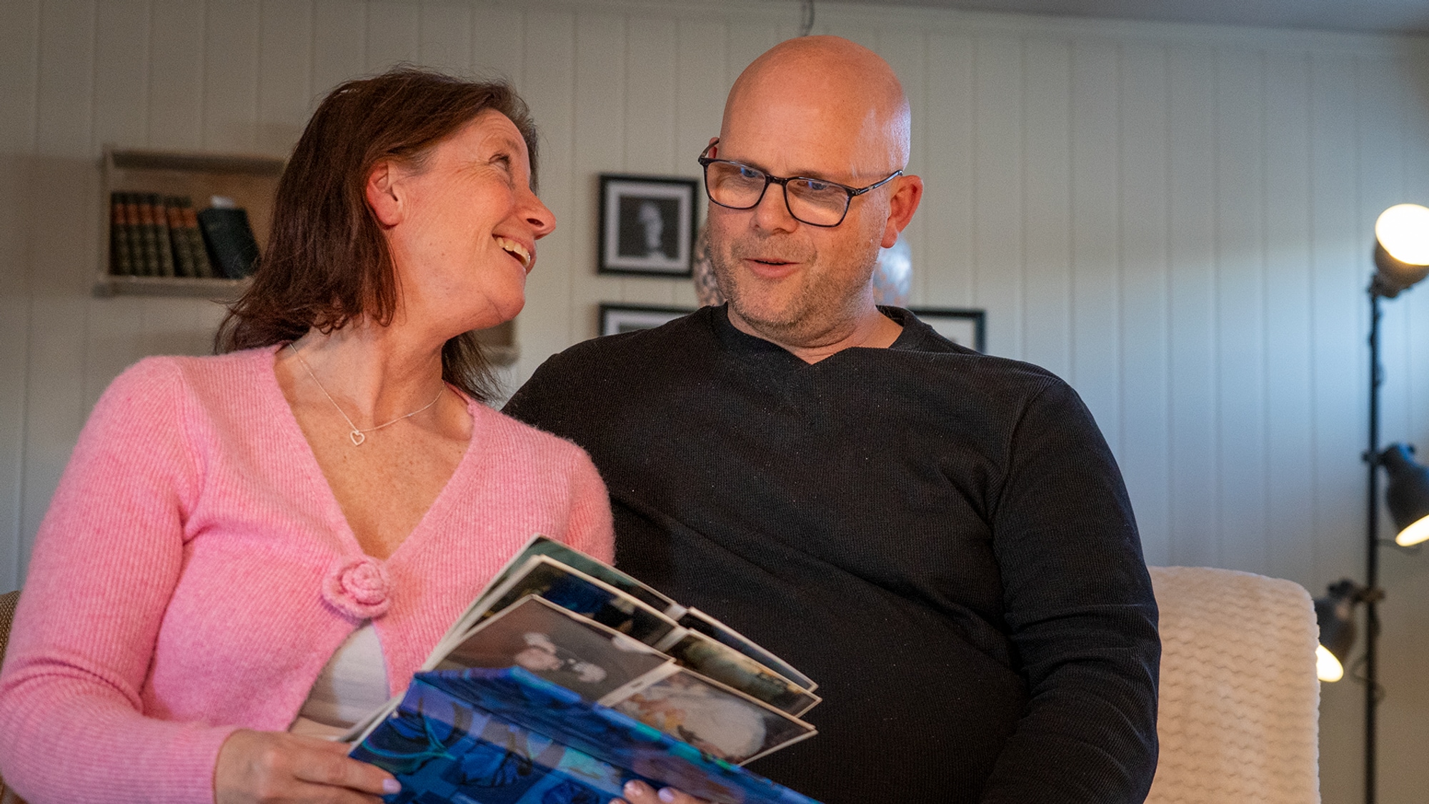 Kvinne med rosa jakke ser smilende opp på mann i svarte klær. Holder barnealbum i hendene. 