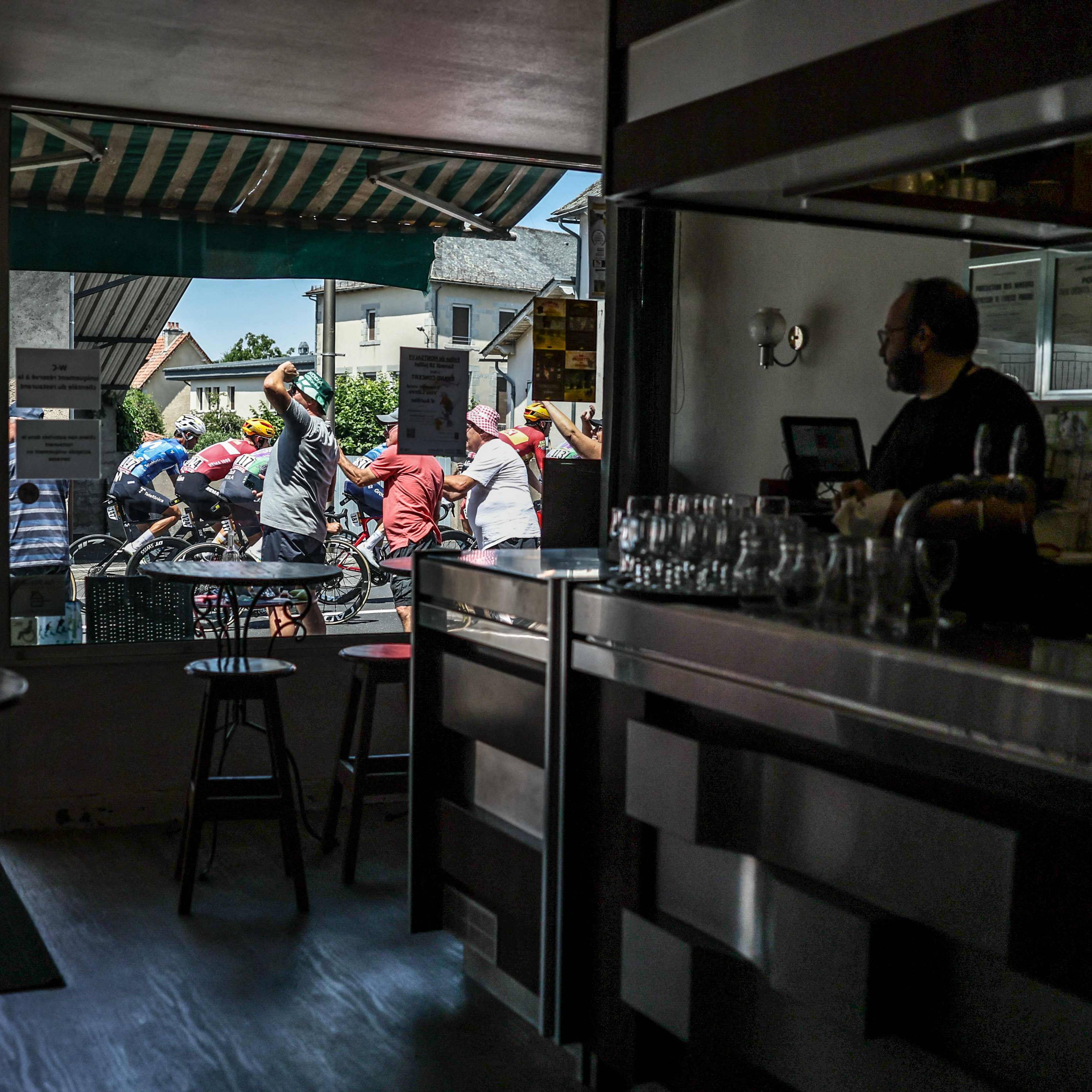 Sykkelritt pågår utenfor en bar, sett gjennom et vindu der en ansatt står ved disken. (Bildebeskrivelsen er laget av en KI-tjeneste)