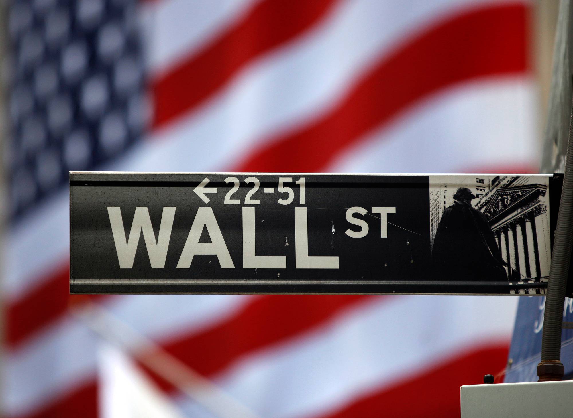 Et Wall Street-skilt er plassert i forgrunnen. I bakgrunnen er et uskarpt amerikansk flagg. Skiltet viser "WALL ST" med piler. Bildet er fotografert i dagtid. (Bildebeskrivelsen er laget av en KI-tjeneste)