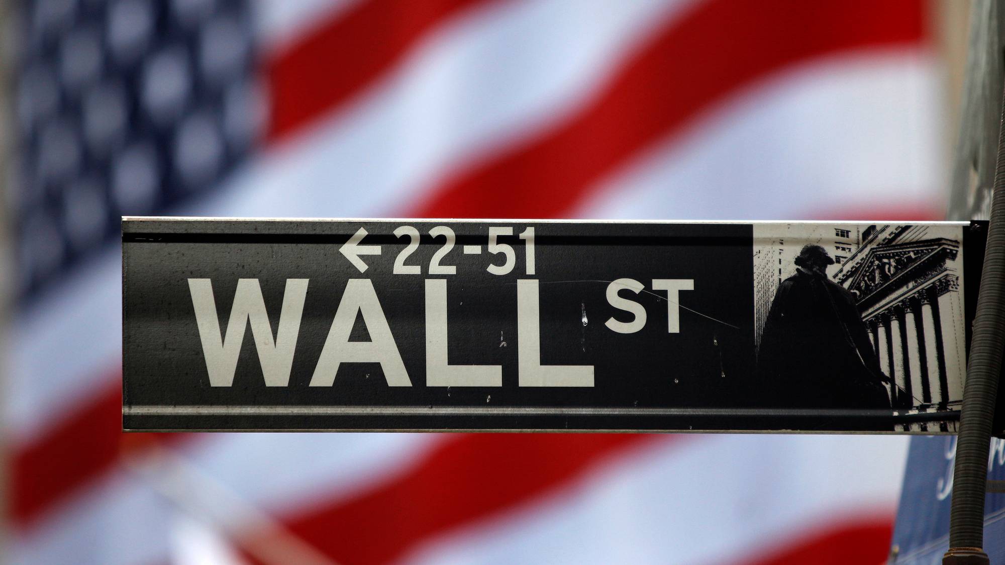 Et Wall Street-skilt er plassert i forgrunnen. I bakgrunnen er et uskarpt amerikansk flagg. Skiltet viser "WALL ST" med piler. Bildet er fotografert i dagtid. (Bildebeskrivelsen er laget av en KI-tjeneste)