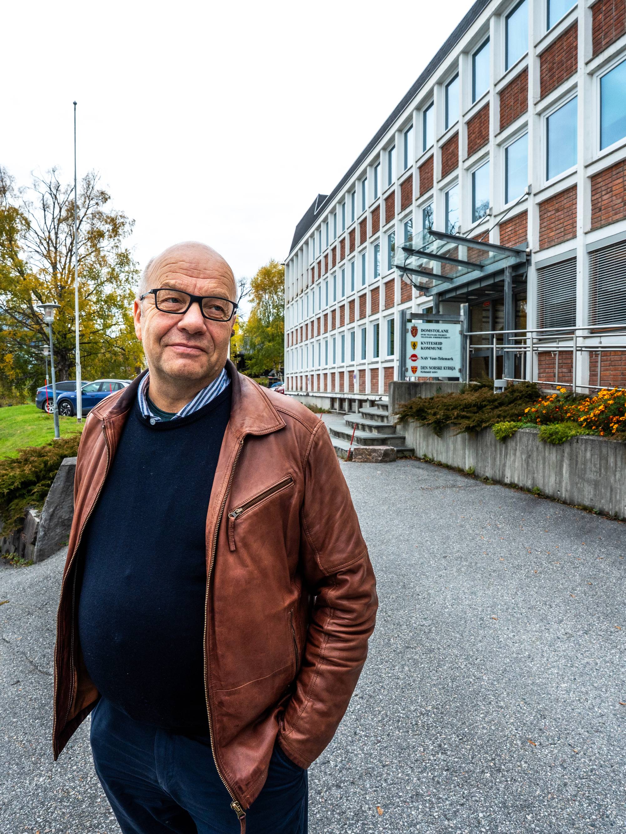 En mann med briller og skinnjakke står foran en offentlig bygning i Kviteseid. (Bildebeskrivelsen er laget av en KI-tjeneste)