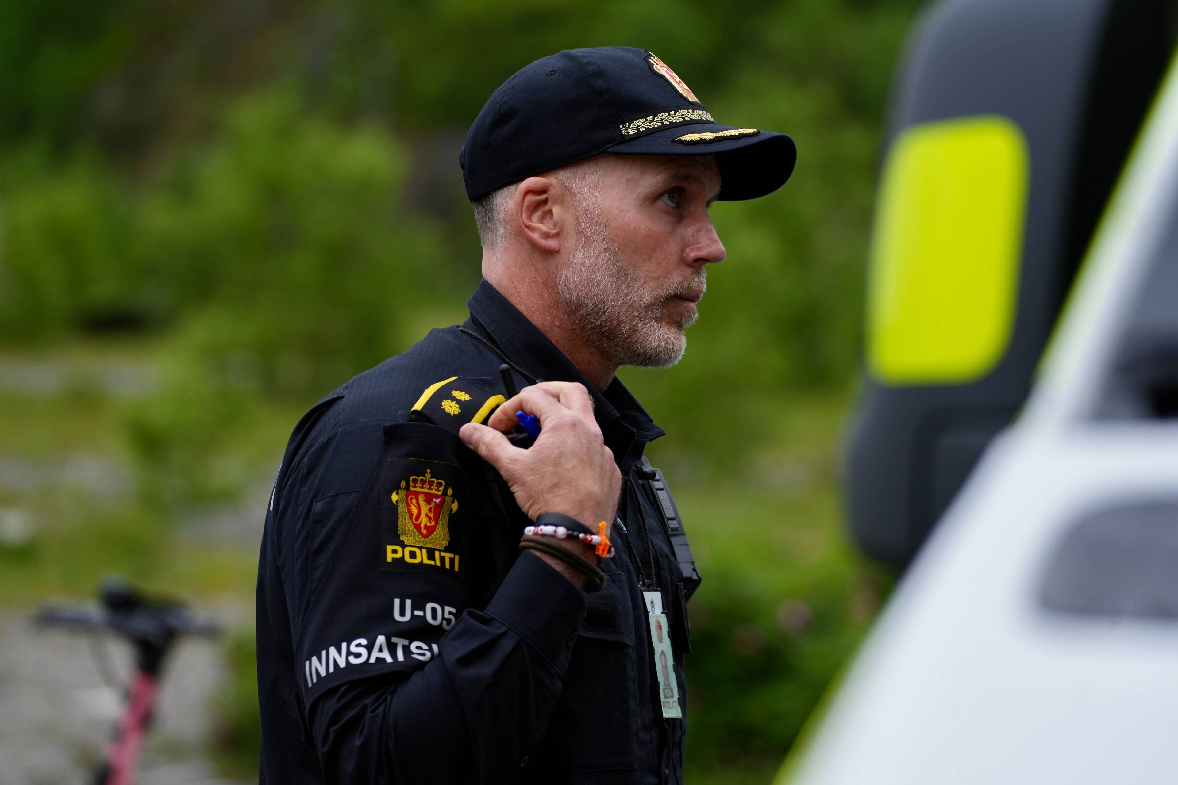 En politimann i uniform står ved siden av et kjøretøy. Han har på seg en caps og har merker på uniformen. Det er grønt landskap i bakgrunnen. (Bildebeskrivelsen er laget av en KI-tjeneste)