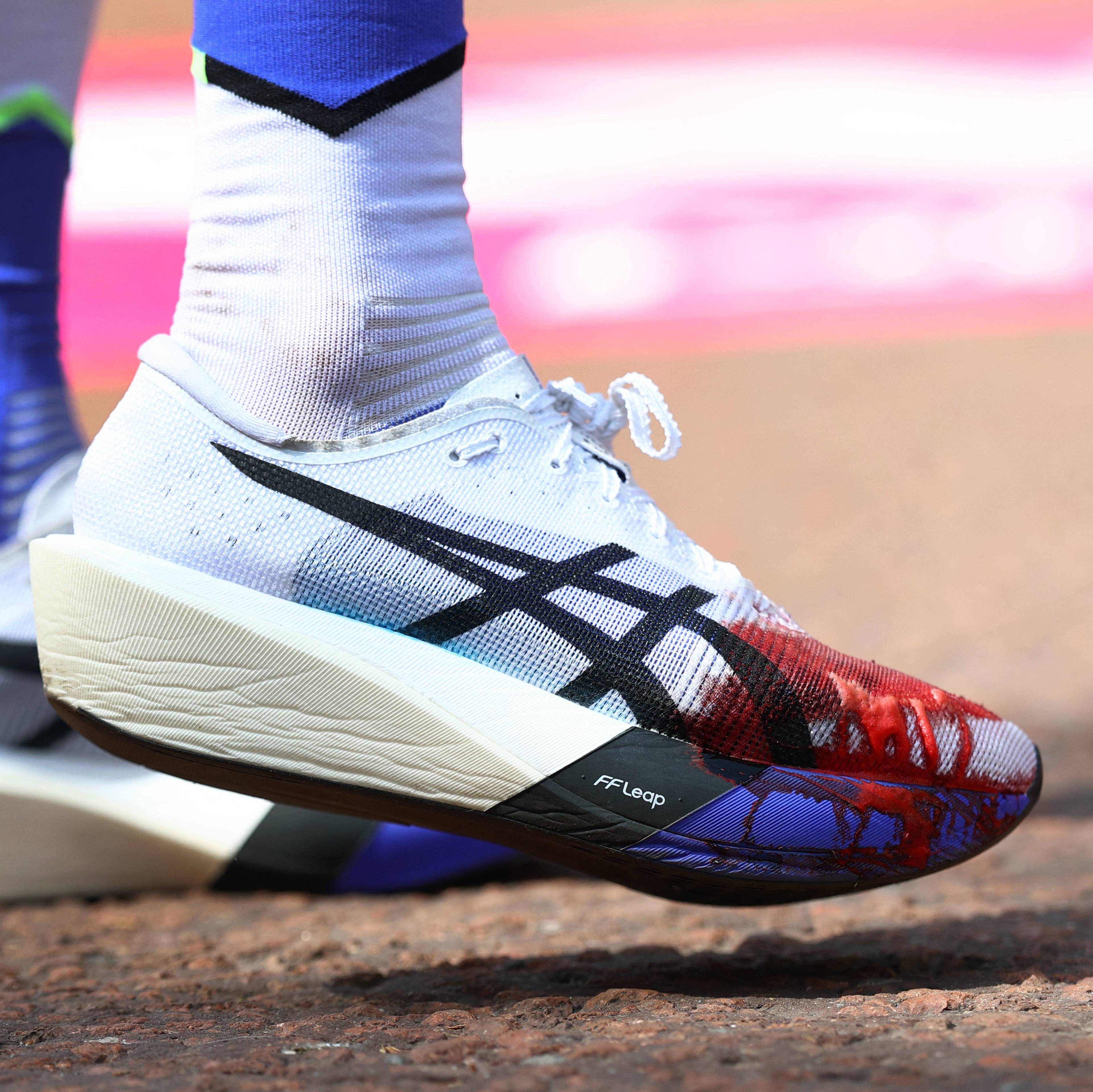 Nærbilde av løpesko og sokker med blå detaljer. Den hvite skoen har et svart Asics-logo og rød og blå print på tåen. Den andre skoen er hvit med svart Asics-logo og en bred, lys såle. En del av en rosa bakgrunn er synlig. (Bildebeskrivelsen er laget av en KI-tjeneste)