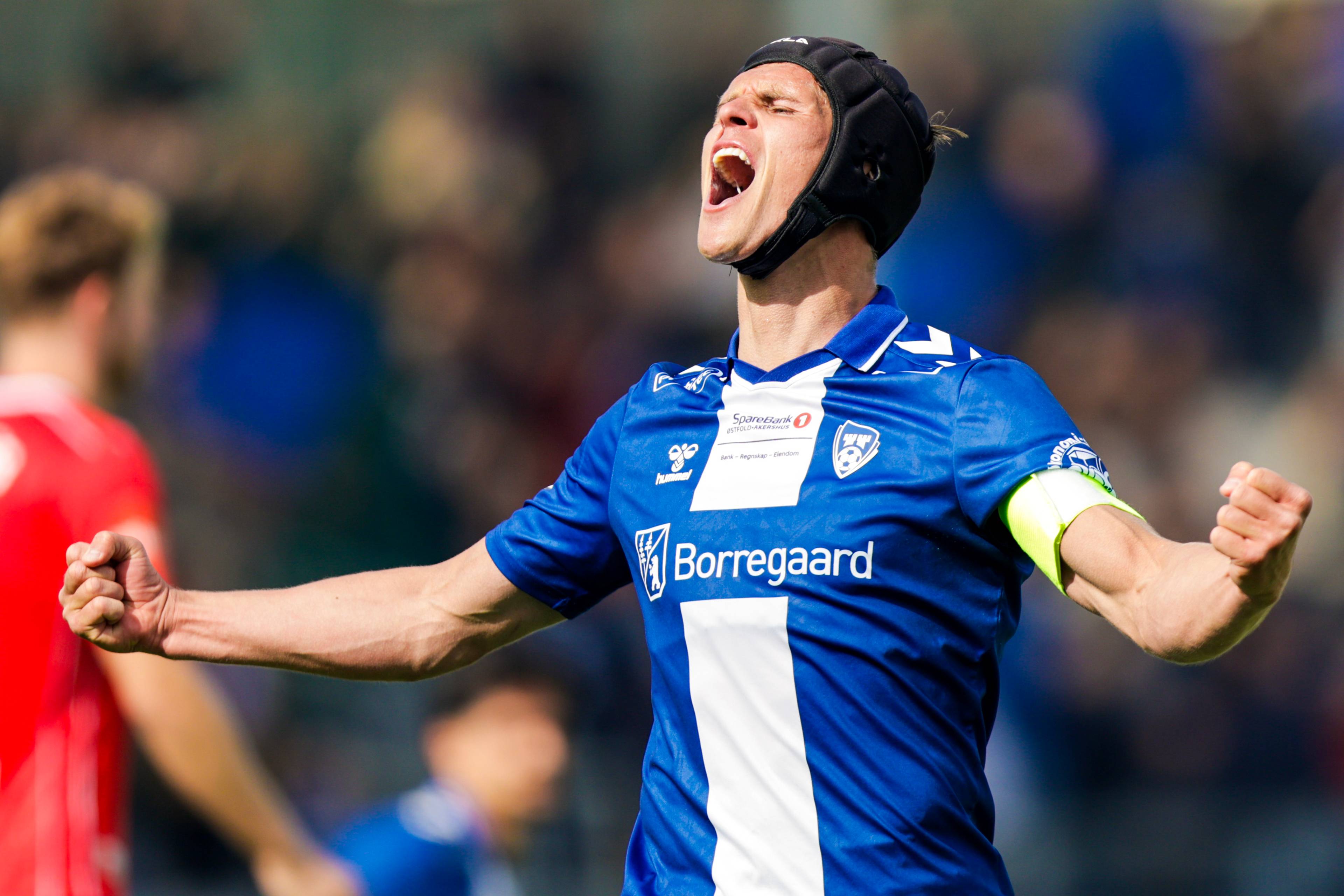 En fotballspiller i blå drakt jubler. Han har på seg en svart hodebeskytter. Spilleren har armene løftet og knyttede never. I bakgrunnen skimtes andre spillere og et publikum. (Bildebeskrivelsen er laget av en KI-tjeneste)
