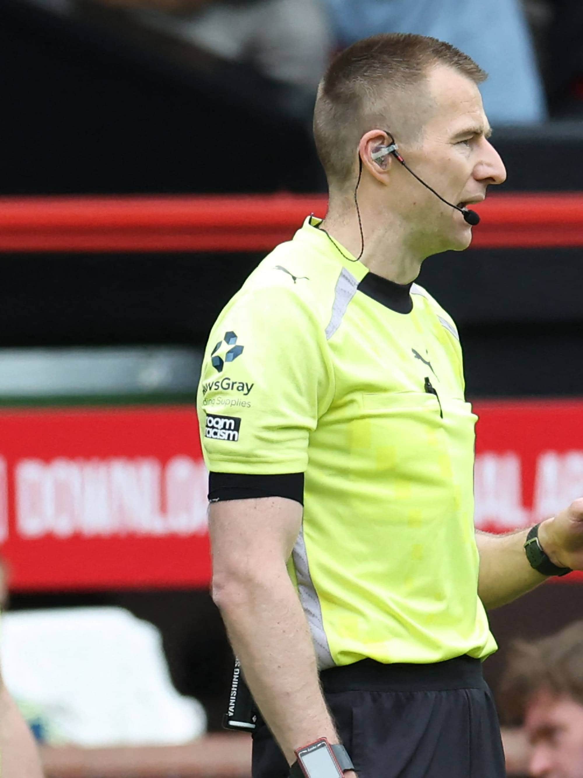 Dommer i gult og svart. Han er på en fotballbane, med røde seter og mursteinsvegger bak seg. Dommeren har en mikrofon ved øret. Bak ham er det andre personer. (Bildebeskrivelsen er laget av en KI-tjeneste)