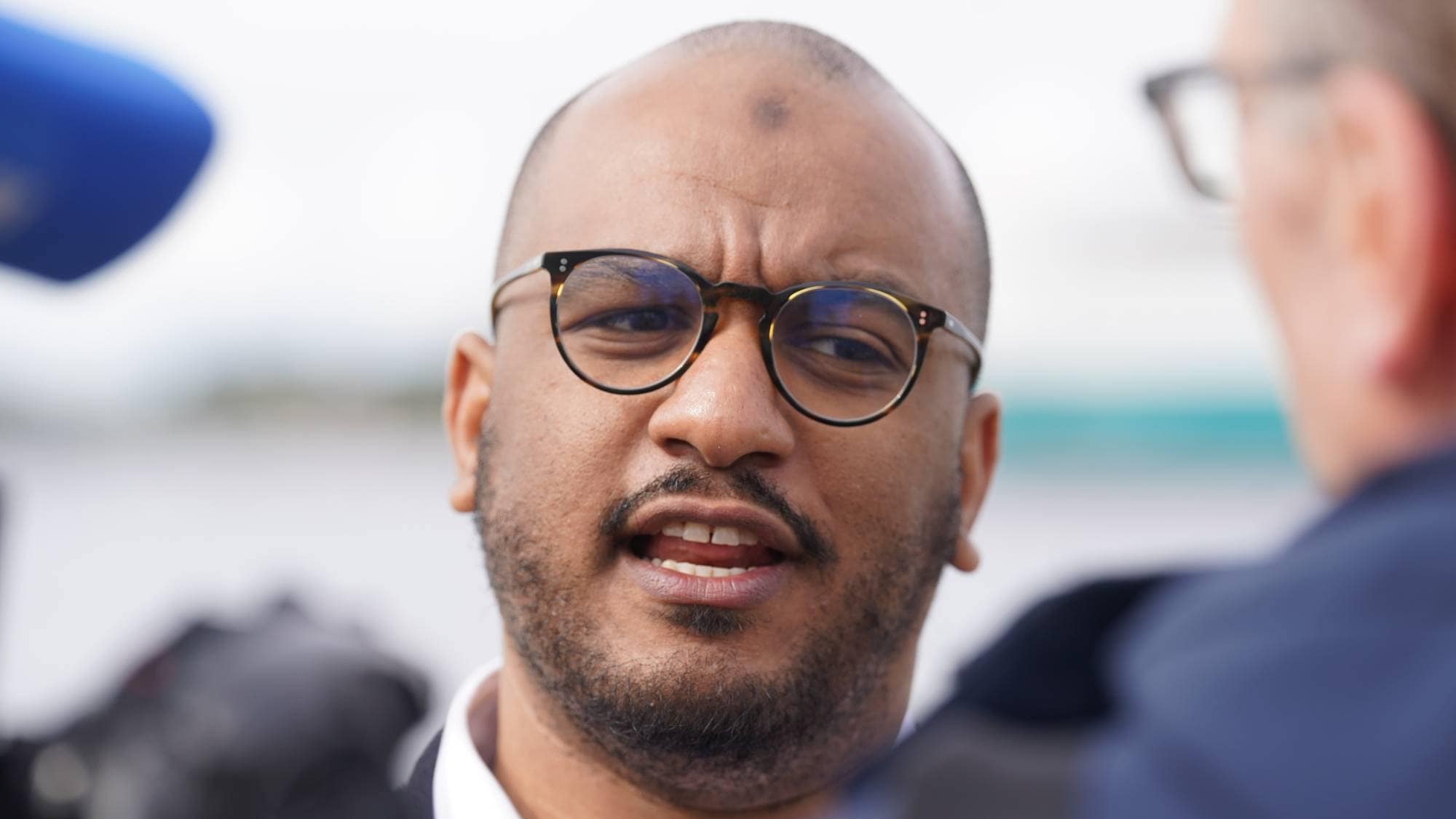 Et nærbilde av en mann med briller som snakker. En mikrofon er synlig til venstre. En annen person er uskarpt synlig til høyre. (Bildebeskrivelsen er laget av en KI-tjeneste)