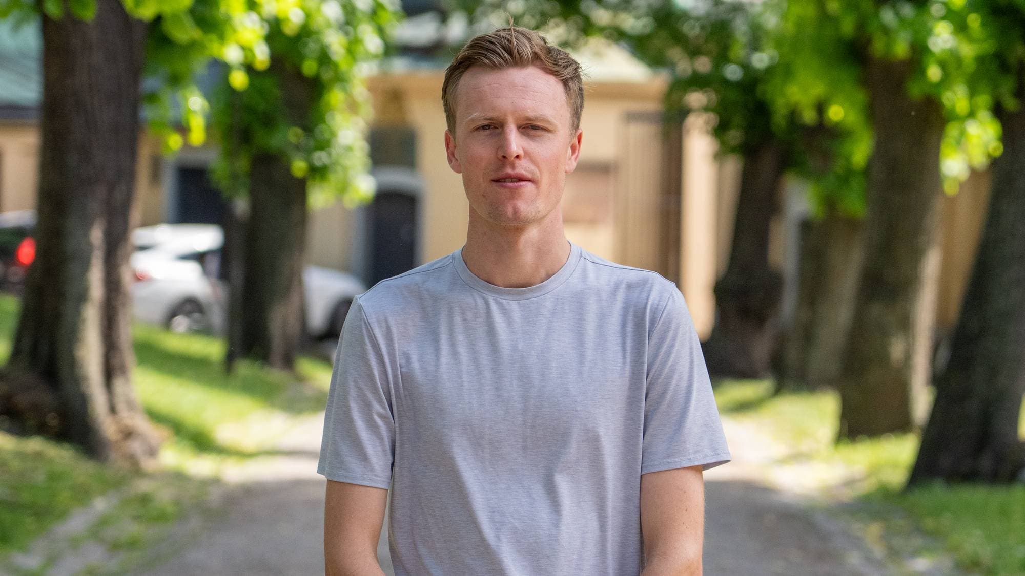 En mann med blondt hår står og ser fremover på en grusvei. Han er kledd i en grå t-skjorte. Bak ham er det trær på begge sider av veien og bygninger. (Bildebeskrivelsen er laget av en KI-tjeneste)