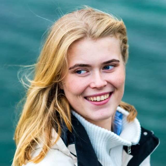 Oda står foran havet i Tromsø. Hun har på seg en varm og vindtett jakke. Hun smiler og ser mot siden mens håret hennes blåser til siden.