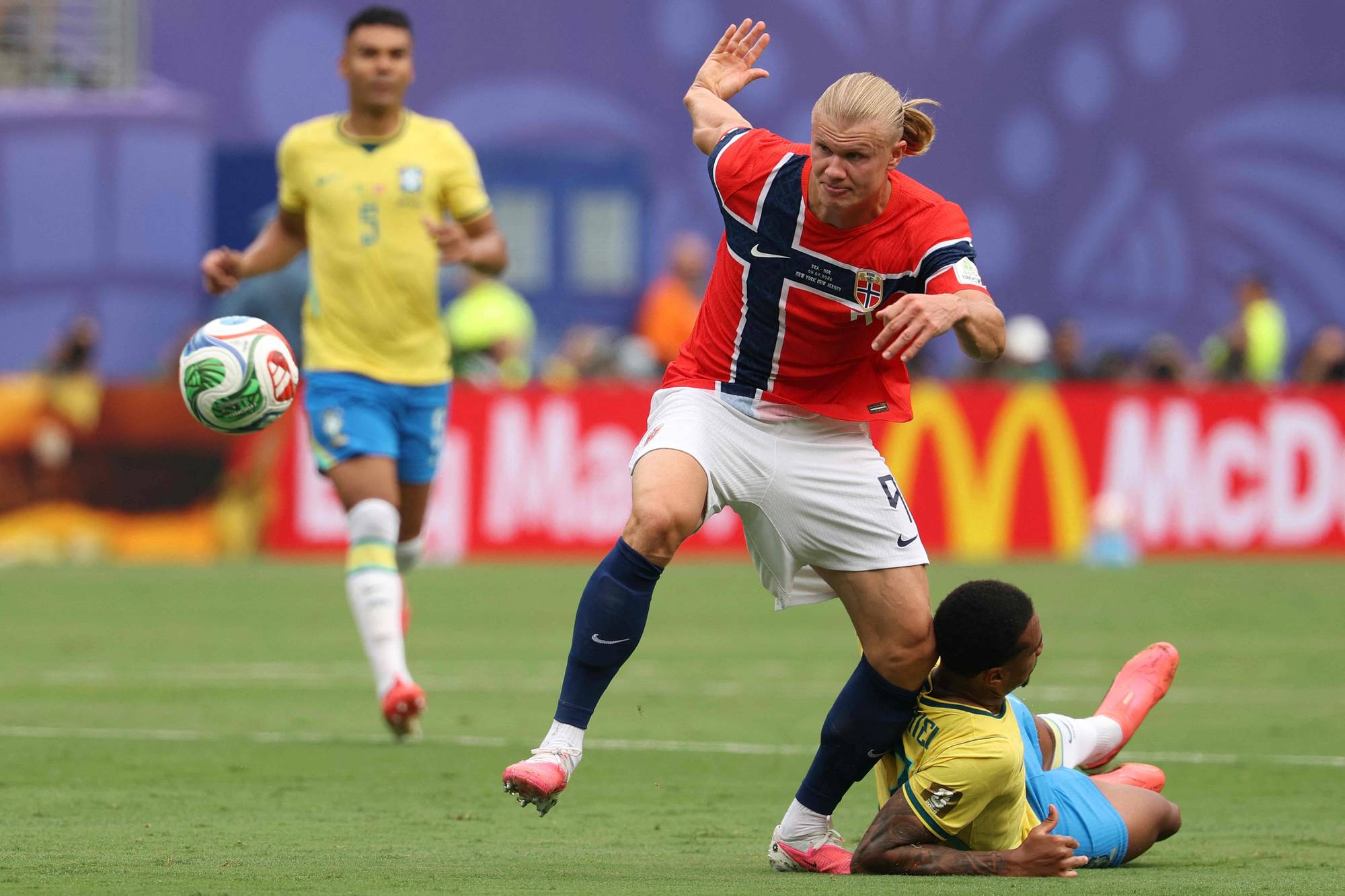 Erling Braut Haaland i norsk landslagsdrakt i duell med en brasiliansk spiller på en fotballbane. (Bildebeskrivelsen er laget av en KI-tjeneste)