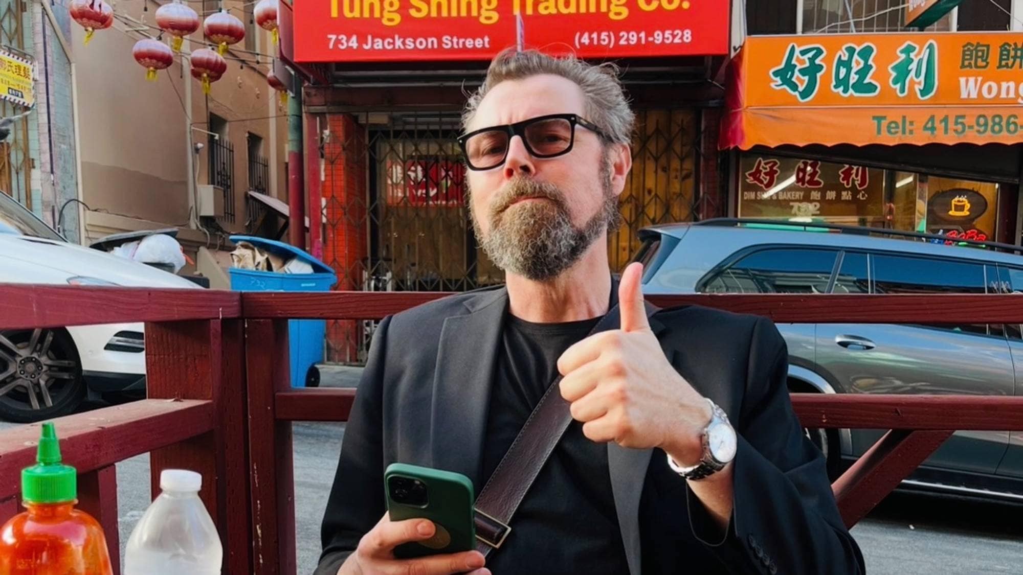 En mann med skjegg og solbriller sitter ved et bord ute. Han holder en telefon i den ene hånden og gir tommelen opp med den andre. Bak ham er en bygate med skilt på kinesisk. (Bildebeskrivelsen er laget av en KI-tjeneste)