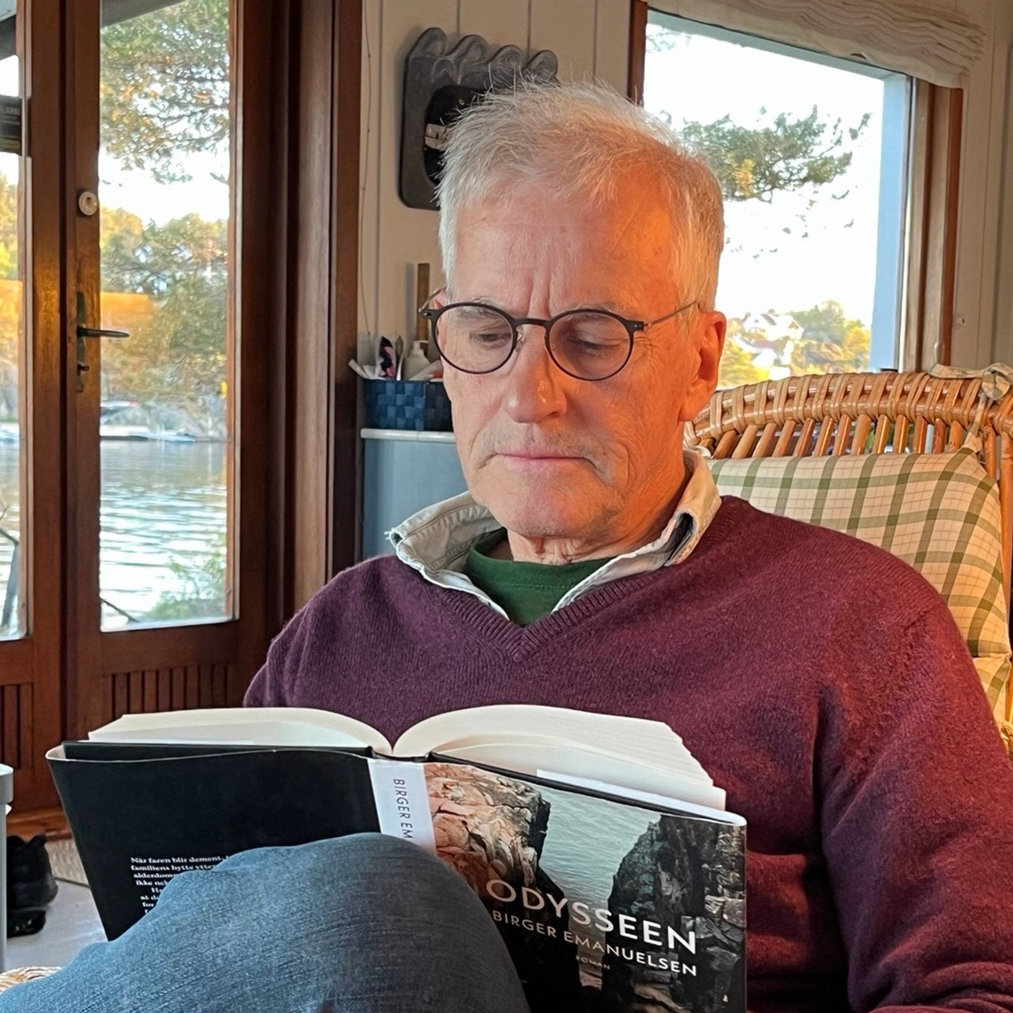 En mann med kort, grått hår (statsminister Jonas Gahr Støre) som sitter i en lenestol og leser boka Odysseen av Birger Emanuelsen, i vinduene skimtes sjø og furutrær.