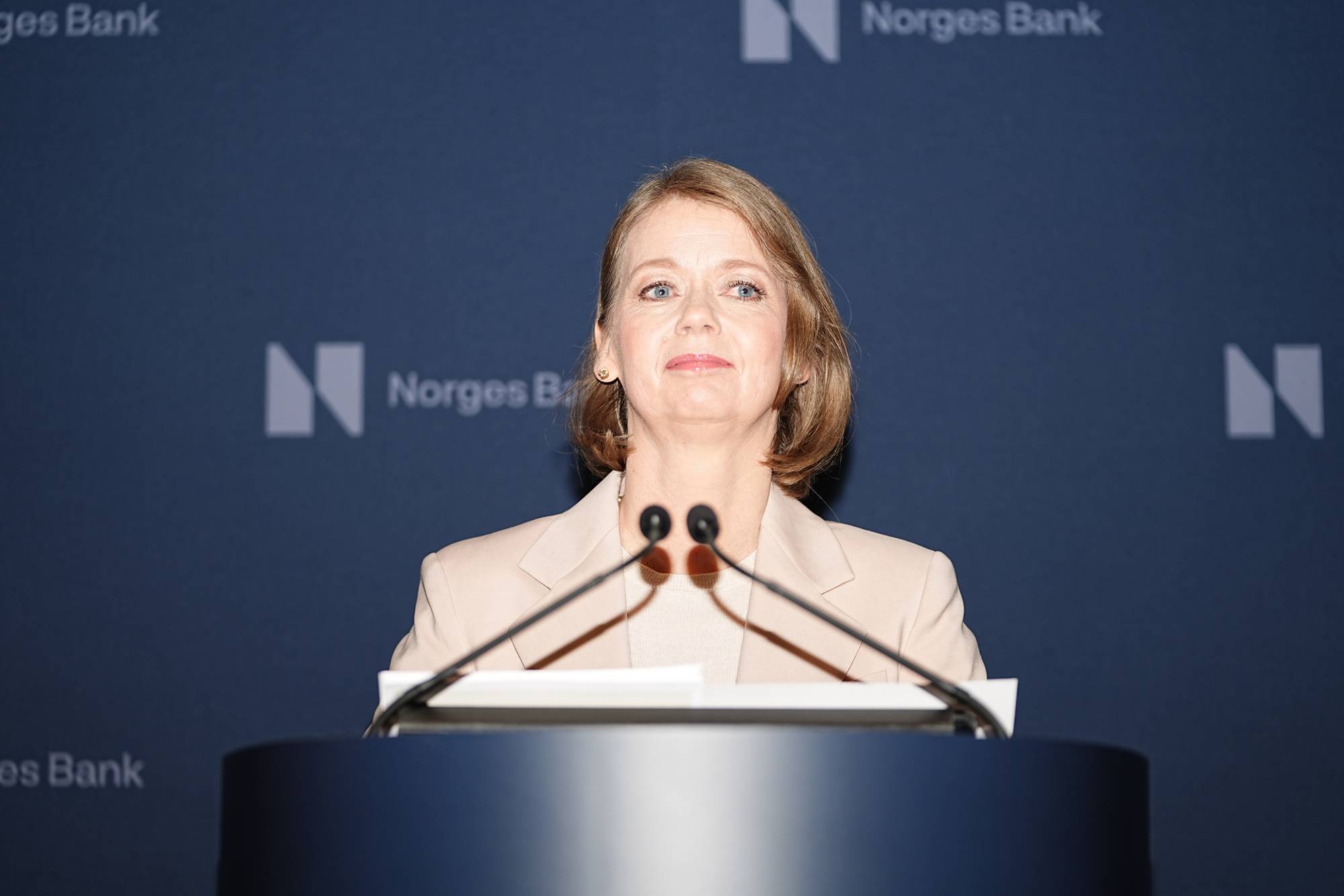sentralbanksjef Ida Wolden Bache står bak et podium med to mikrofoner foran seg. Hun ser mot høyre og smiler. Bak henne er en mørkeblå bakgrunn med logoen til Norges Bank. (Bildebeskrivelsen er laget av en KI-tjeneste)