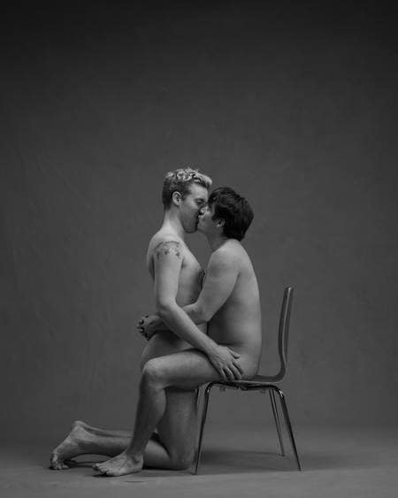 En naken mann med mørkt hår sitter på en stol mens han kysser en lyshåret naken mann som står på kne mellom bena hans