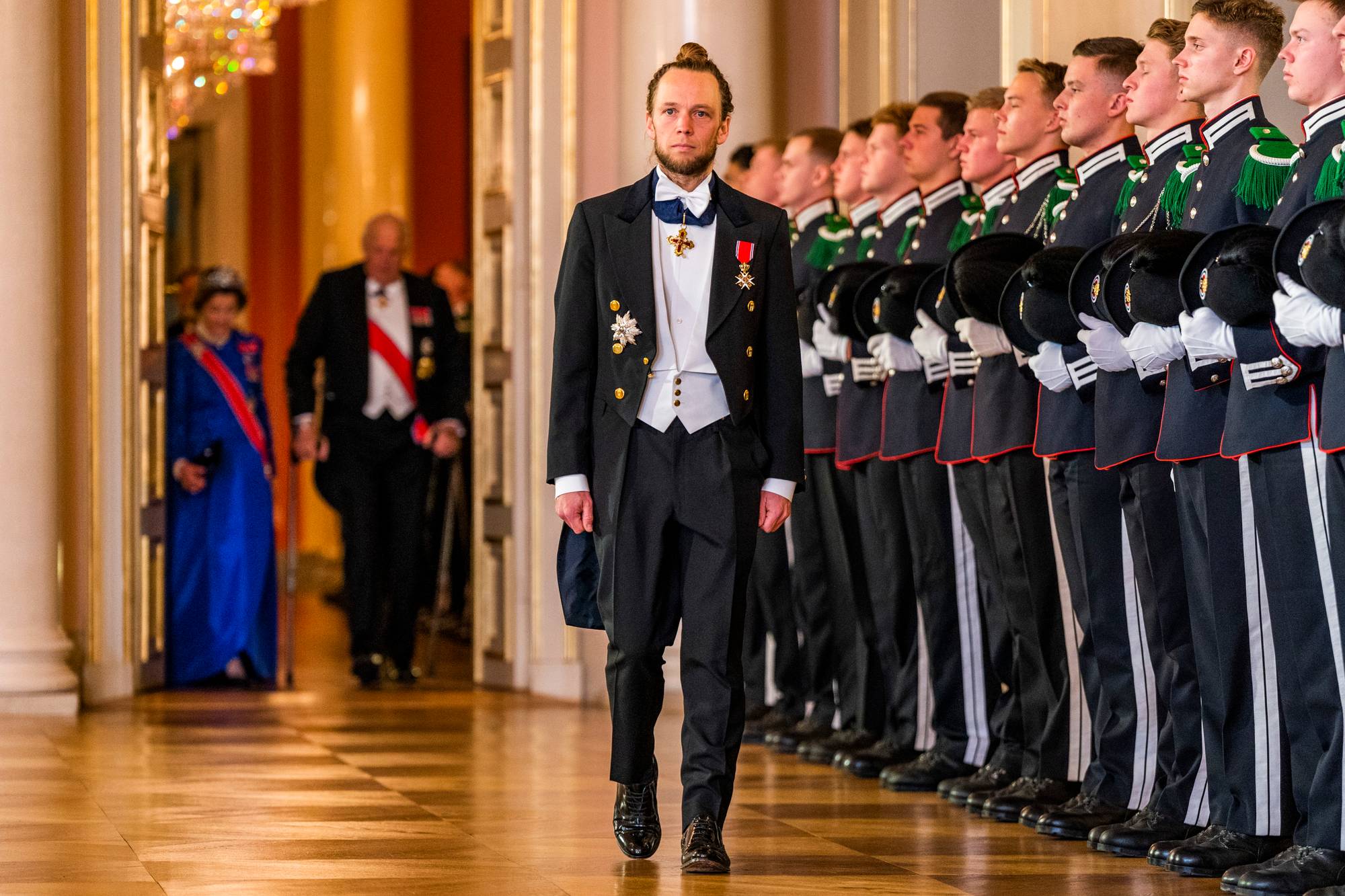 Hoffsjef Olav Heian-Engdal går i prosesjon i Det kongelige slott. Bak ham kommer dronning Sonja og kong Harald. En rekke militære uniformerte står på siden. (Bildebeskrivelsen er laget av en KI-tjeneste)
