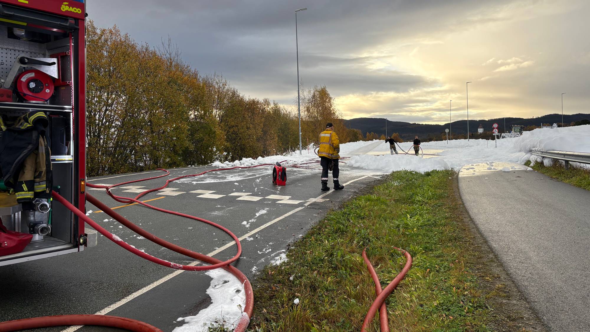 Brannmannskaper driver slukningsarbeid med skum på en vei ved siden av en brannbil. (Bildebeskrivelsen er laget av en KI-tjeneste)