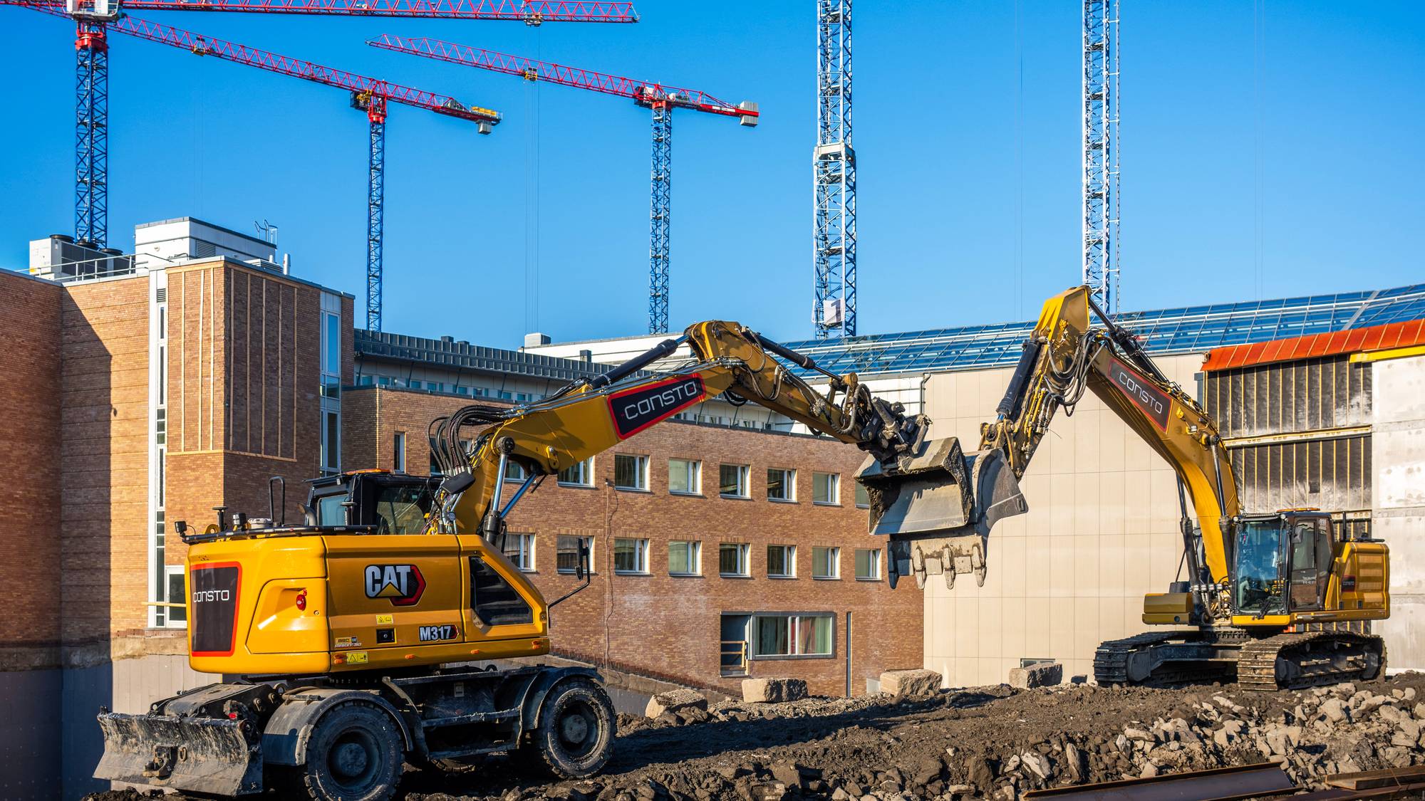 To gule gravemaskiner er i arbeid på en byggeplass. De river en mursteinsbygning. Tre byggekraner står i bakgrunnen. (Bildebeskrivelsen er laget av en KI-tjeneste)