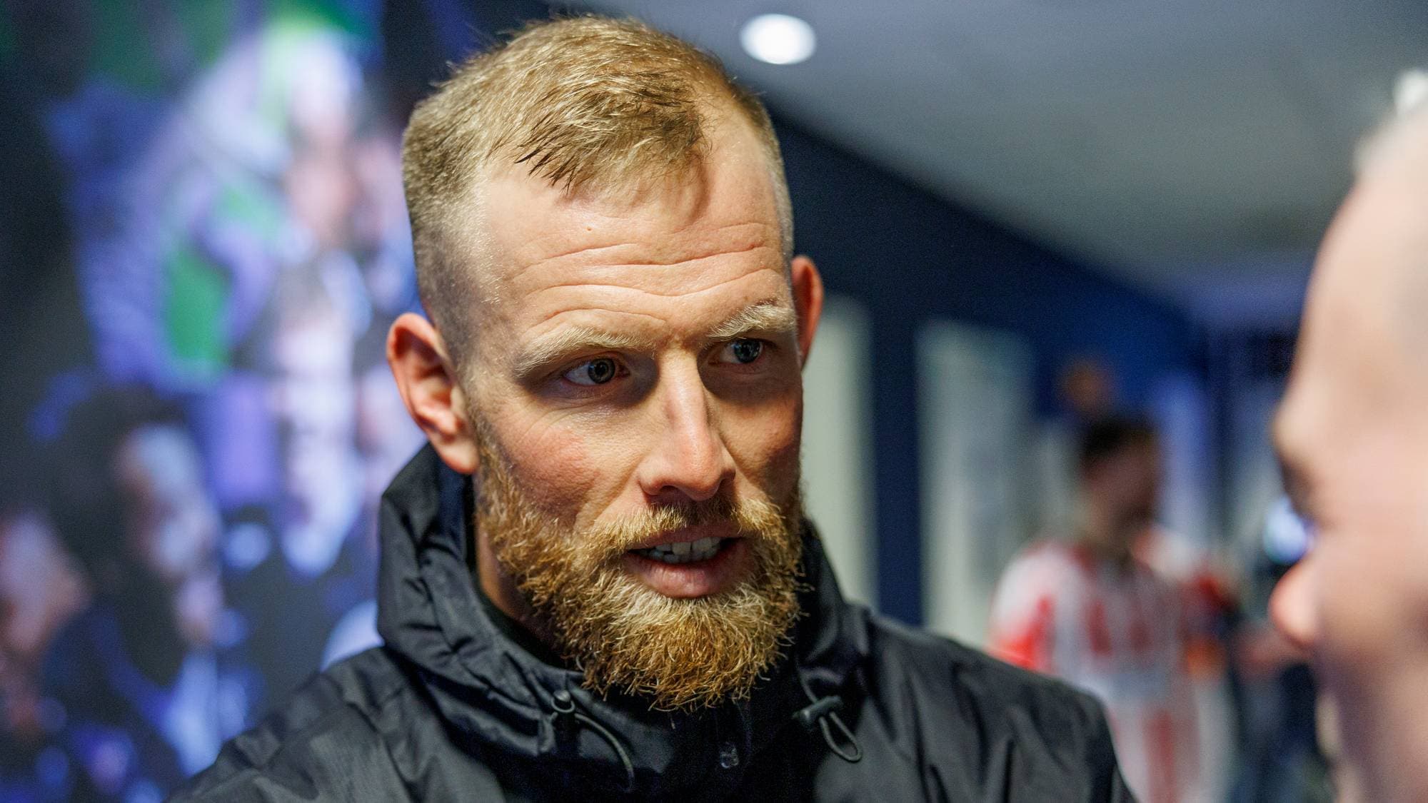 En mann i svart jakke står og blir intervjuet av en journalist med mikrofon. Bakgrunnen viser en gruppe mennesker i utydelige former, som deltar i en sportsrelatert aktivitet. Intervjusituasjonen skjer i et lukket miljø, muligens en arena eller et studio. Mannen ser alvorlig ut mens han lytter til spørsmålene som stilles.