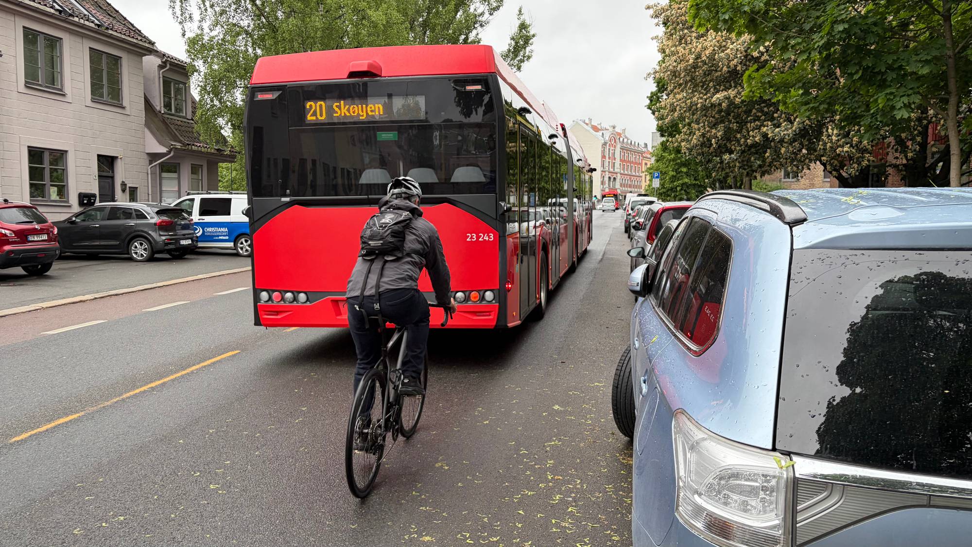 En syklist sykler bak en rød buss på en bygate. Bussen har rutenummer 20 og viser destinasjonen Skøyen. Flere biler er parkert langs gaten. (Bildebeskrivelsen er laget av en KI-tjeneste)