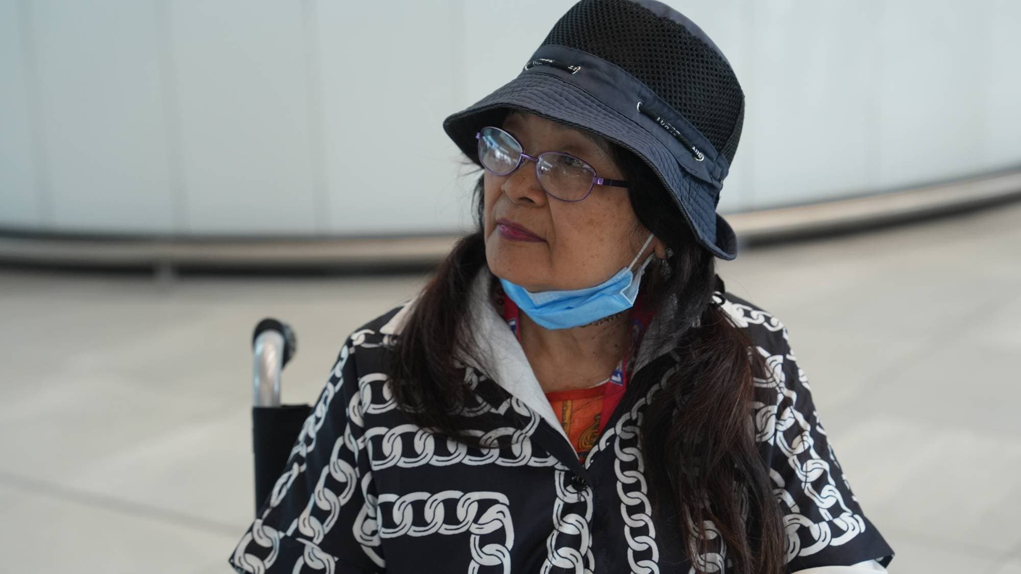 Kvinne med hatt, briller og munnbind sitter i rullestol. Hun ser til siden. (Bildebeskrivelsen er laget av en KI-tjeneste)
